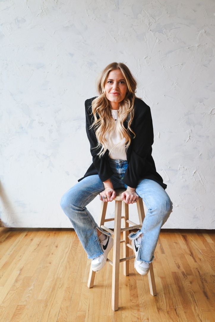 Person wearing blazer and jeans sits casually on wooden stool against white wall.