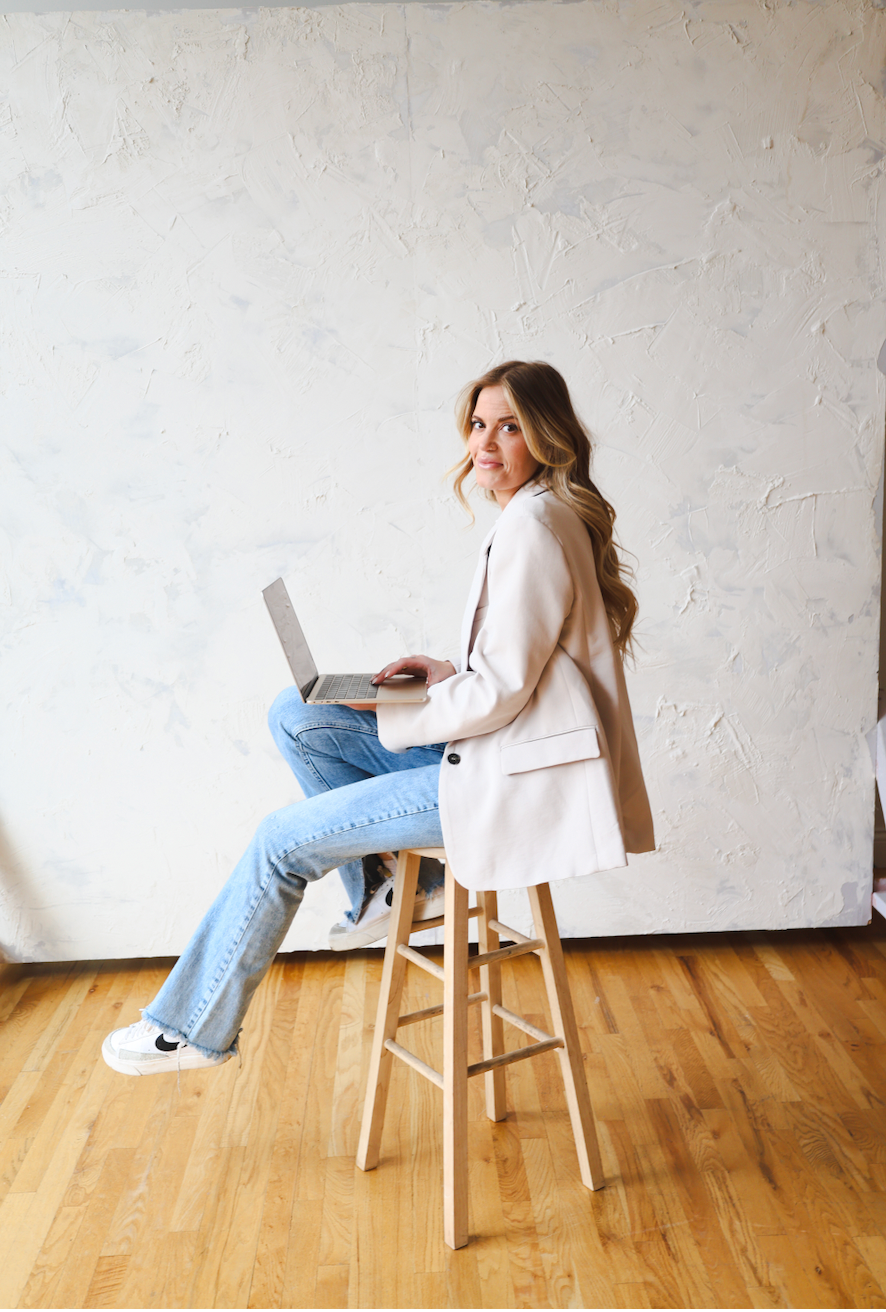Person in light colored top and jeans works on laptop while sitting on wooden stool.