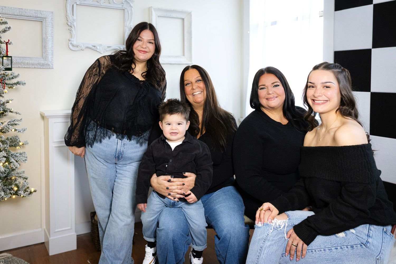 Family portrait session with coordinated outfits in front of checkered wall and Christmas tree.