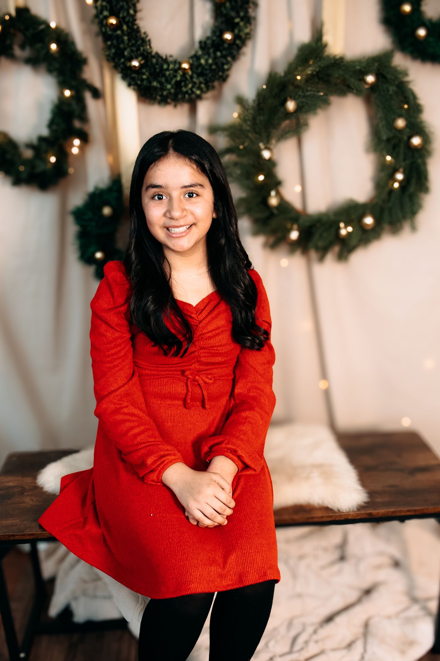 Person in red sweater sitting and smiling in front of Christmas wreaths and twinkling lights.
