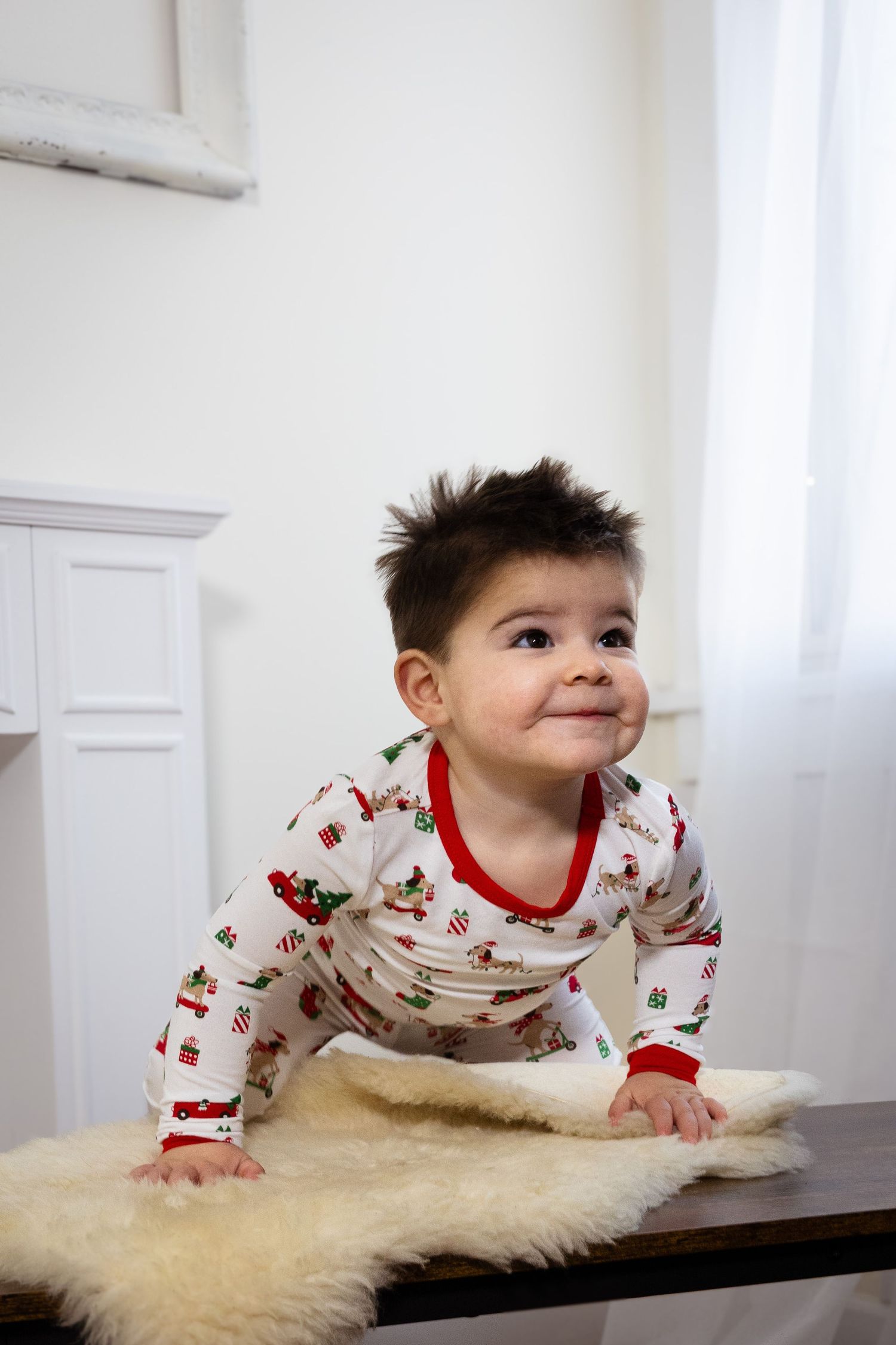 Child in festive pajamas crawls on furry white rug near bright window.