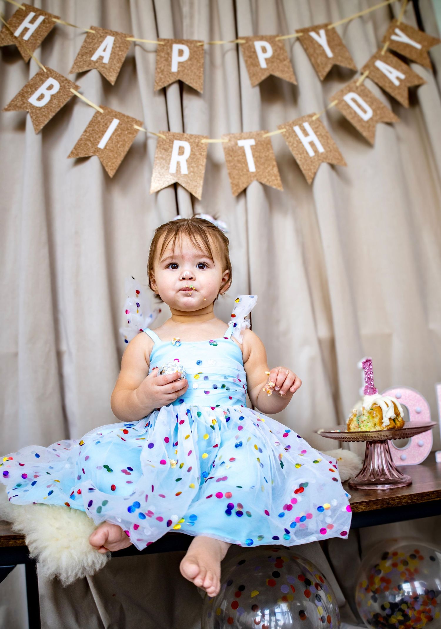 Birthday photo series of baby in blue dress with confetti pattern under burlap banner.