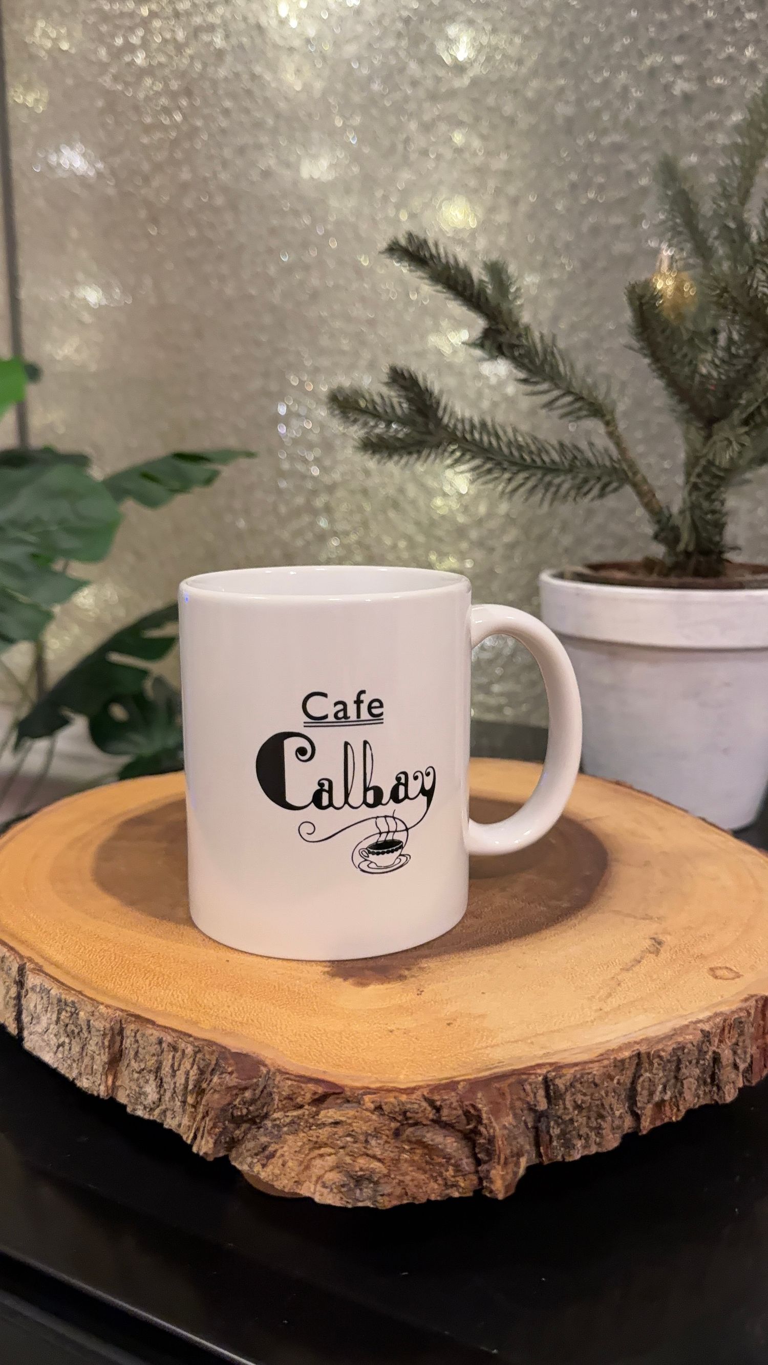 White ceramic coffee cups with cafe logo displayed on natural wood slice with evergreen sprigs.