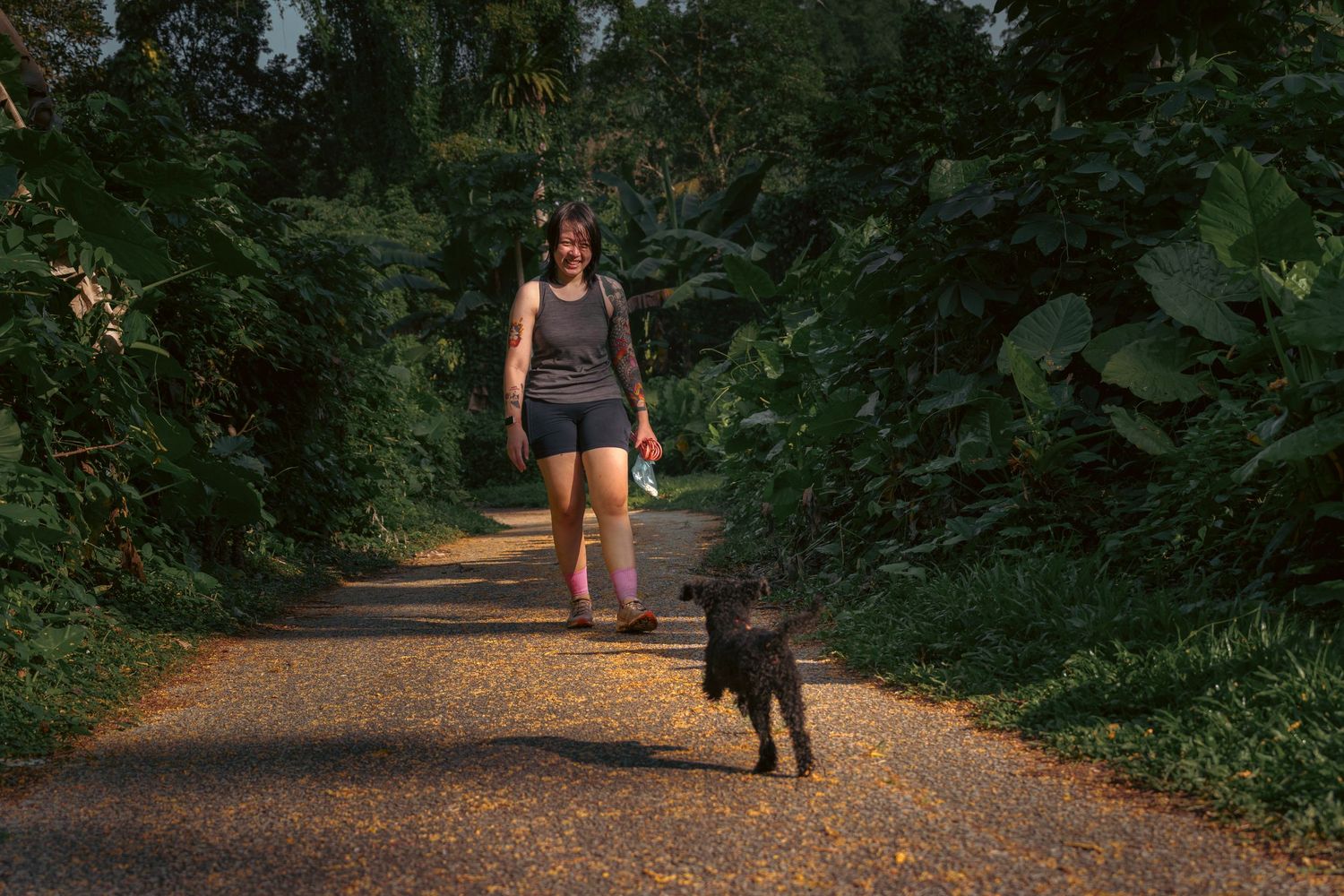 A dirt path through lush green forest with a smiling lady in athletic wear playing with her small dog at Bukit Brown Singapore