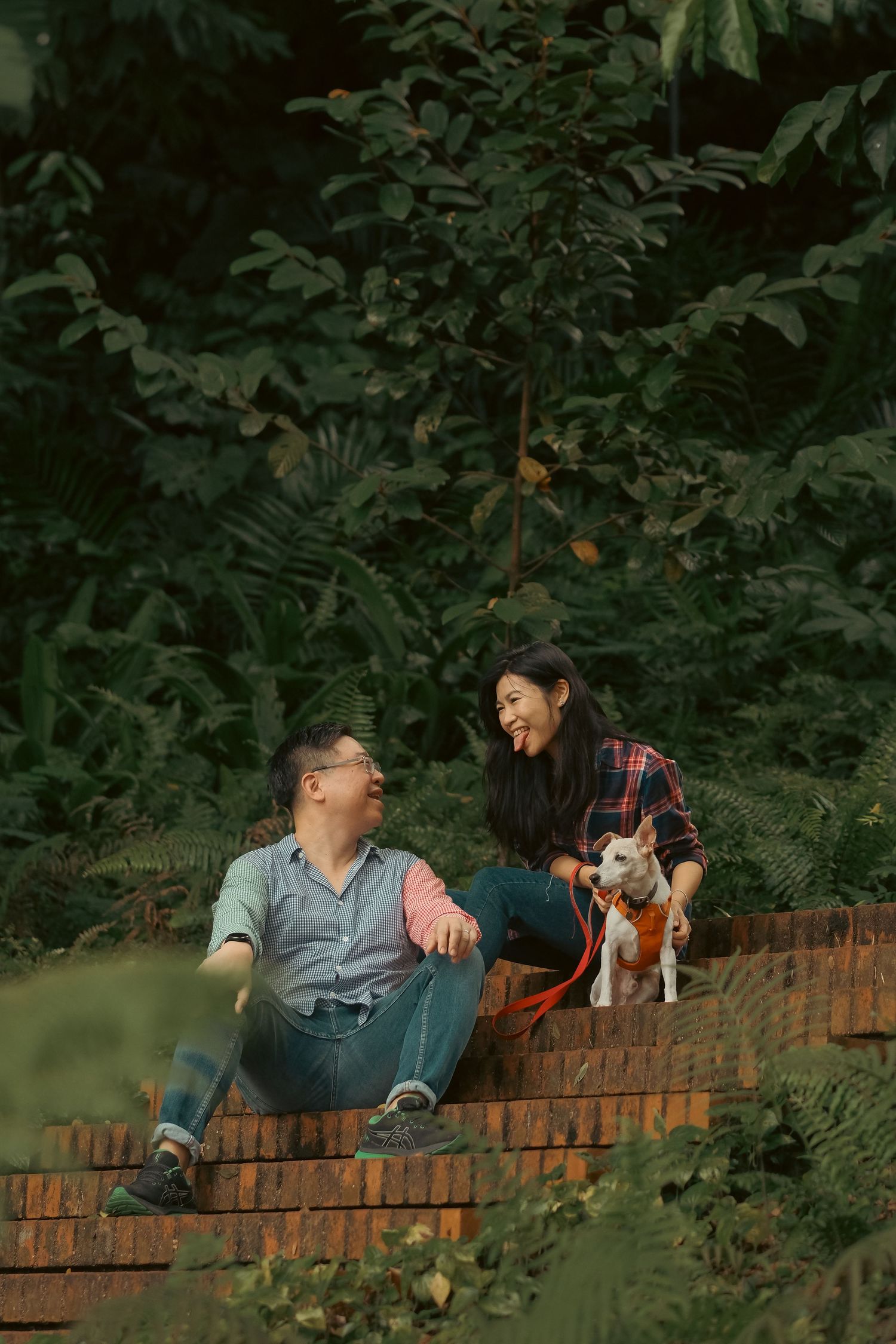 A couple relaxes with their small white dog on a brick wall in a garden setting.