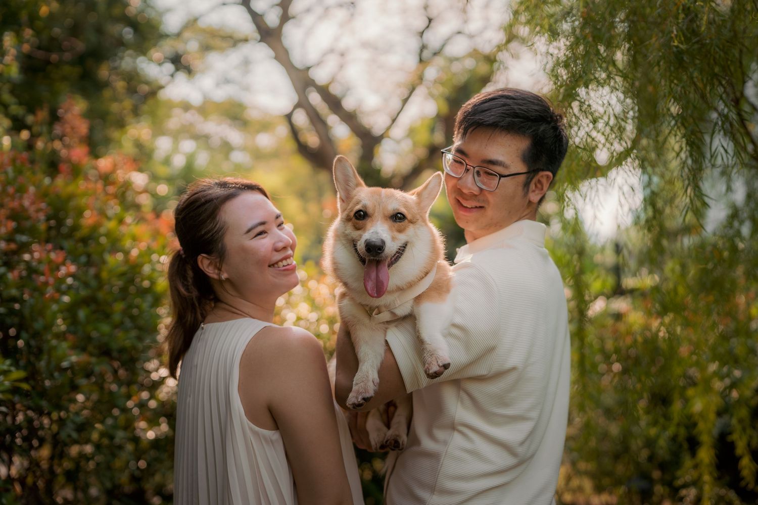 Happy couple holding a Corgi dog outdoors in a garden with colorful foliage backdrop.
