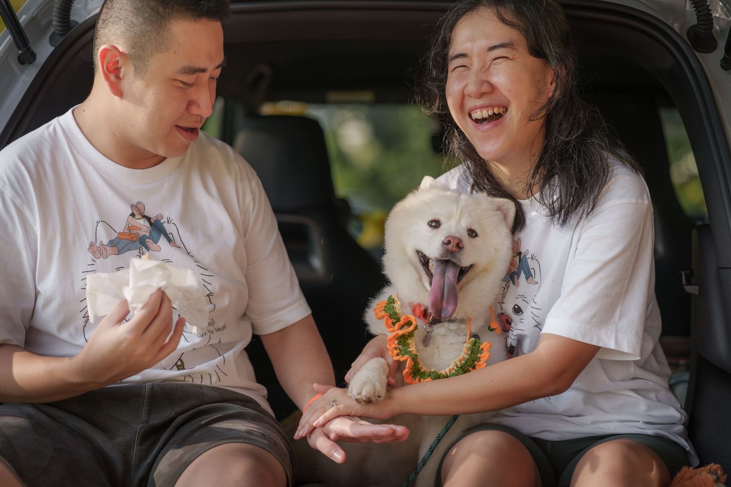 A couple and their small white dog share joyful moments in the back of their vehicle.