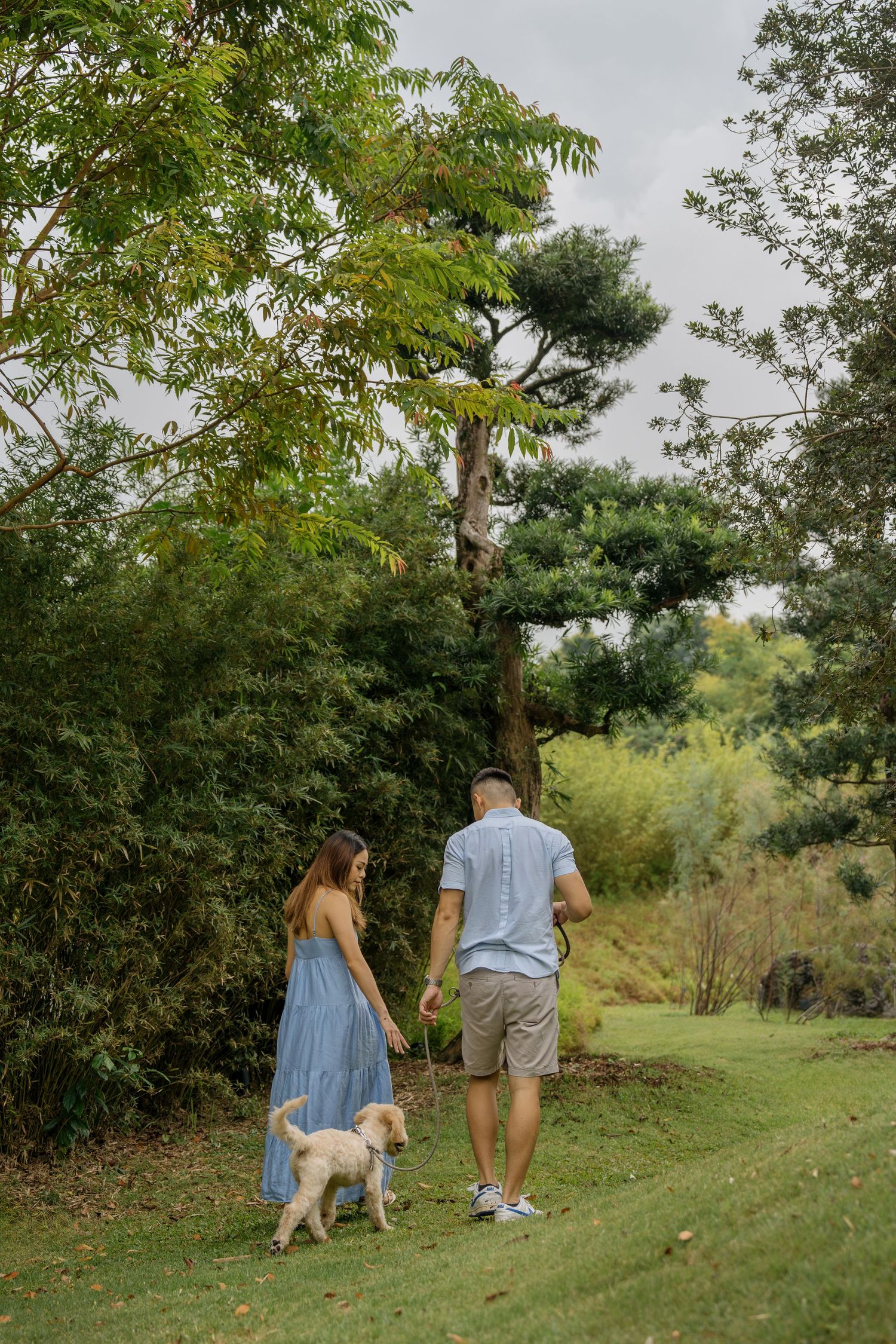 A couple and their fluffy white dog enjoy a stroll through a lush green park on a cloudy day.