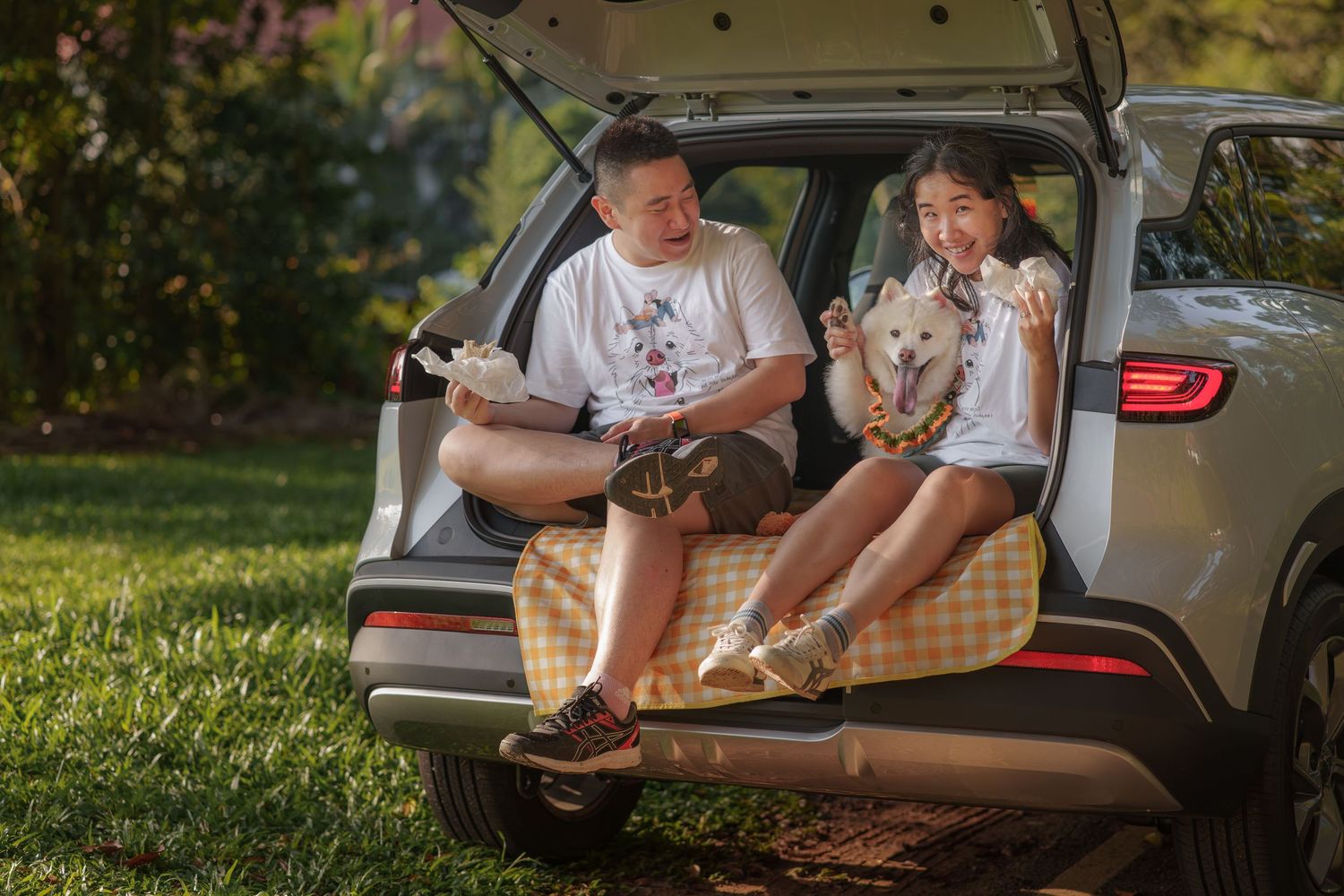 A relaxed couple sitting in their car trunk with their pets during a sunny afternoon.