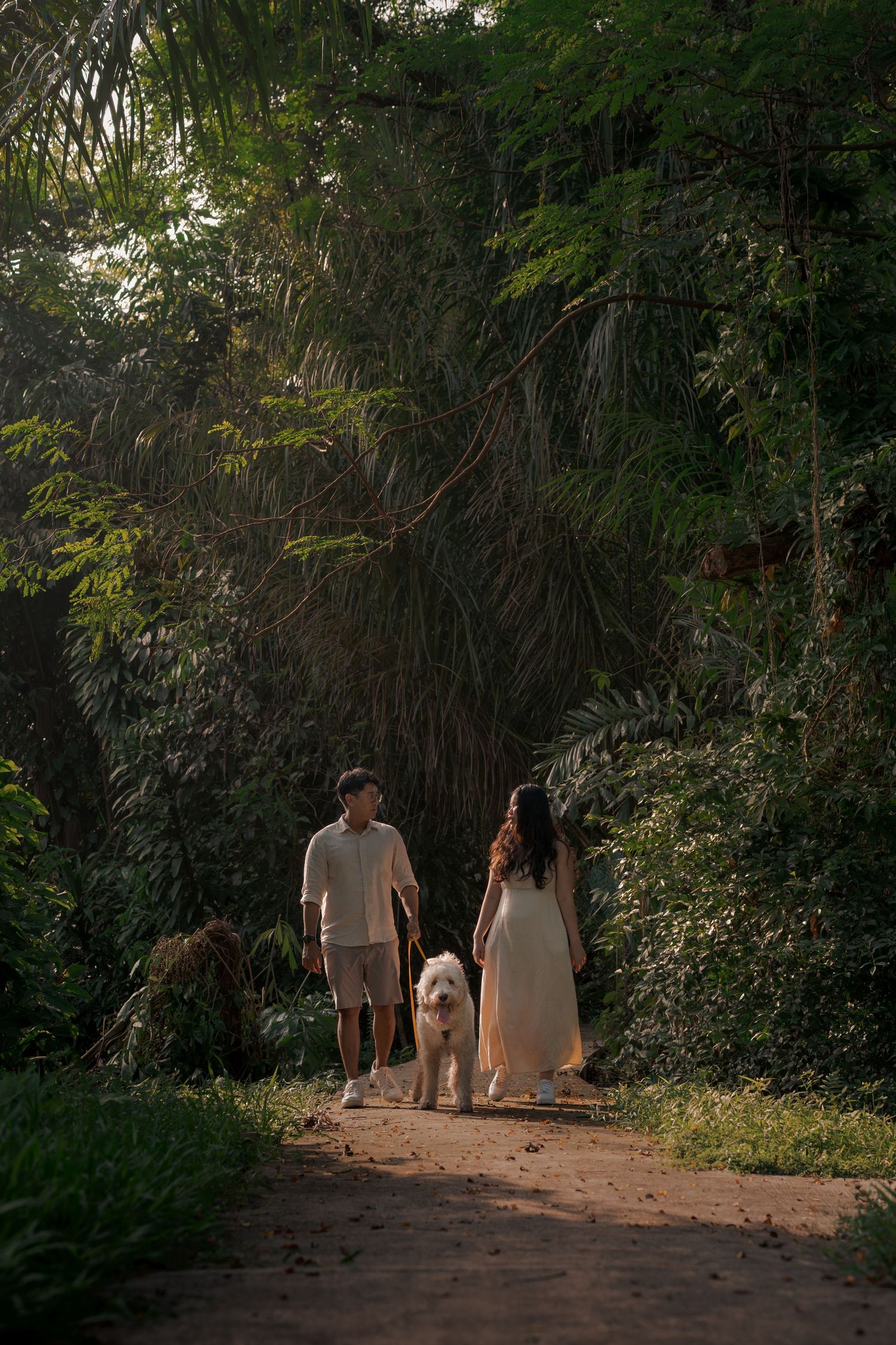 Couple walking white fluffy dog on a serene forest path surrounded by lush tropical greenery.