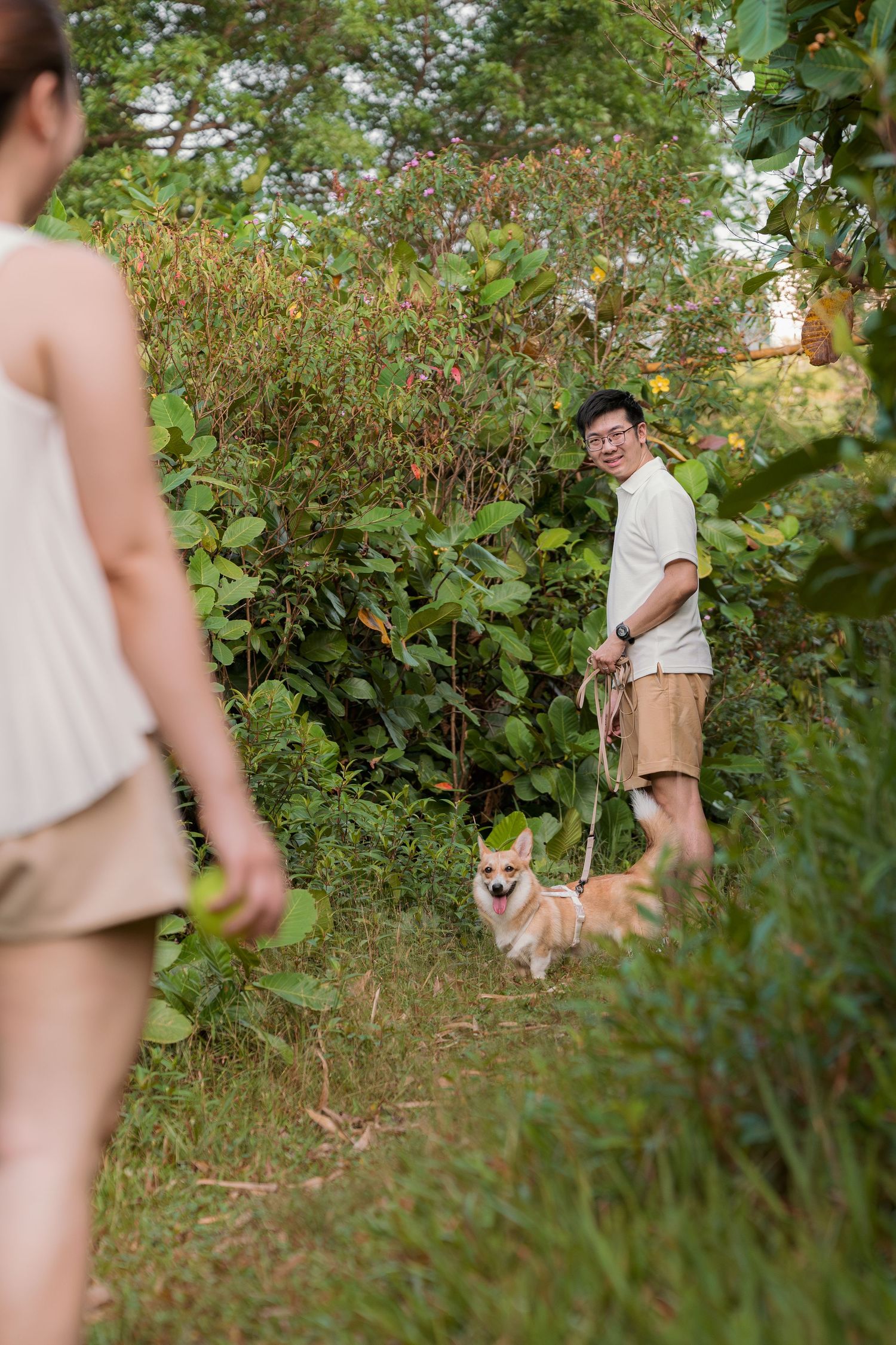 Man walking a Corgi dog on a leash through a lush green trail, smiling at a woman in the foreground.