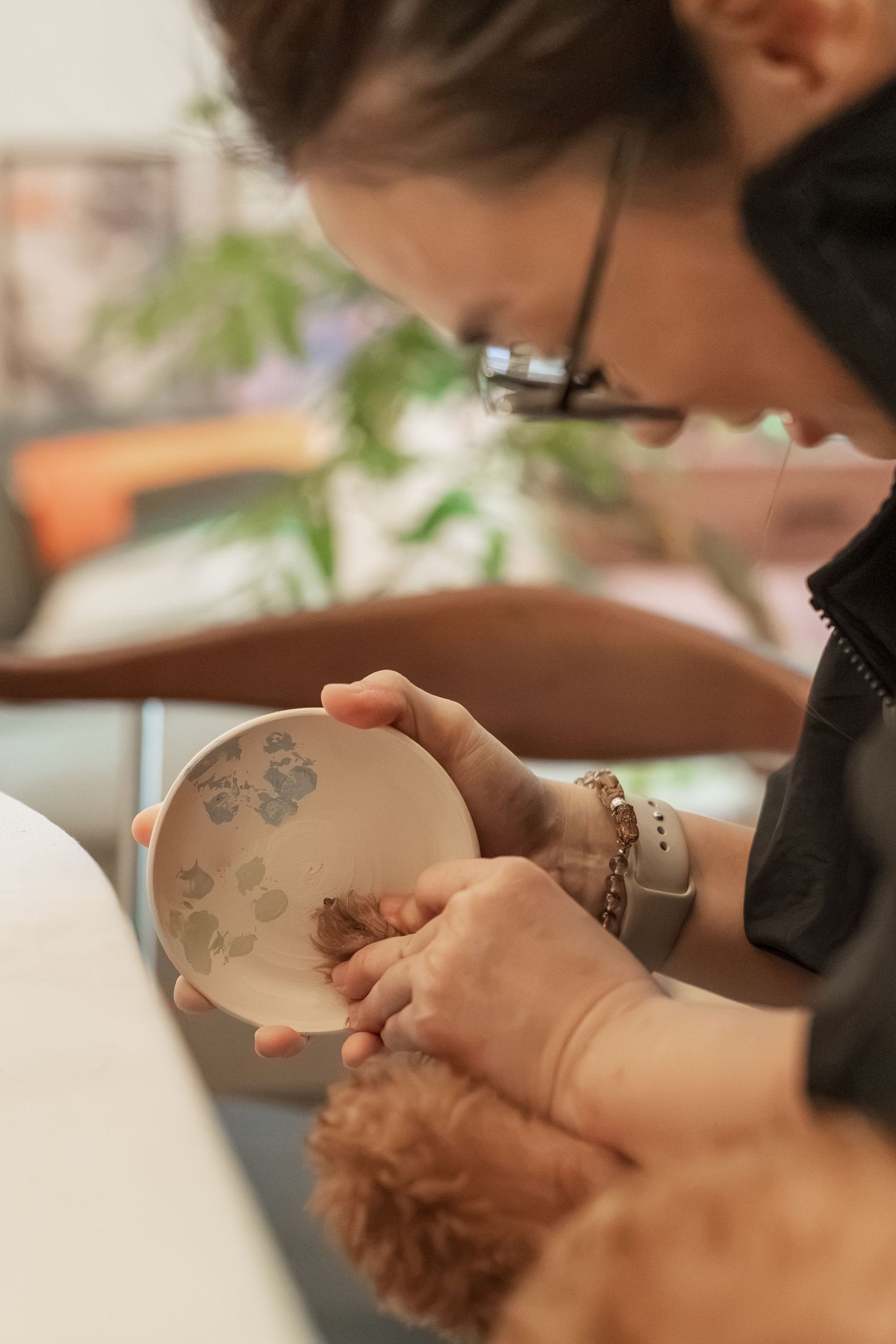 Woman with glasses examining a decorative plate with floral pattern while holding a small curly-haired dog.