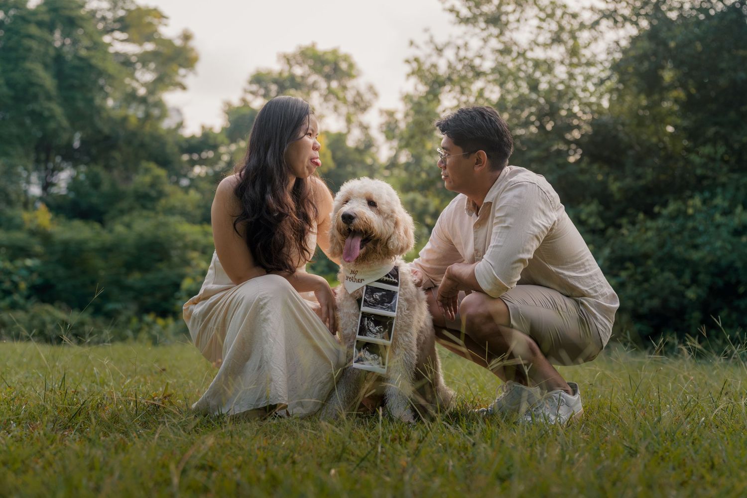 Couple crouching on grass with golden doodle dog holding ultrasound photos, pregnancy announcement in park.