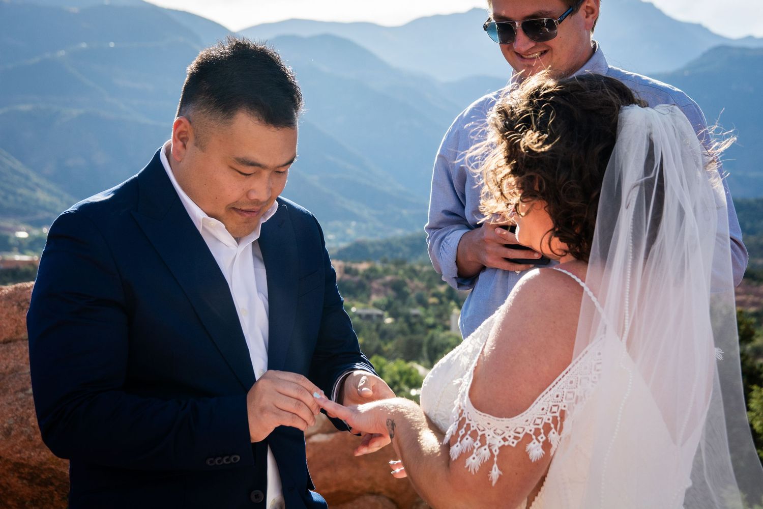 Groom placing wedding ring on bride's finger during outdoor ceremony with mountain backdrop.