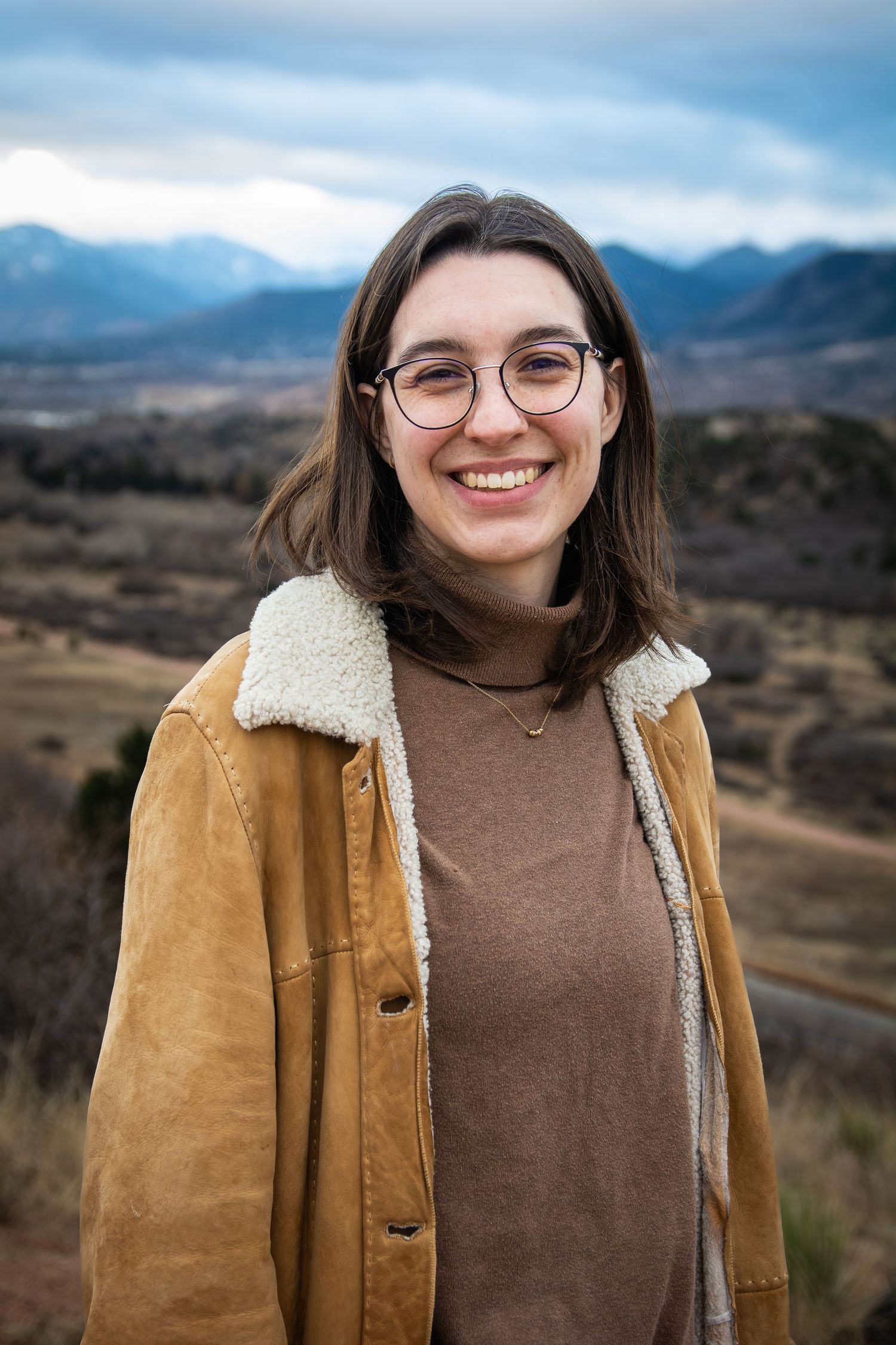 Smiling young woman in sherpa jacket posing outdoors with mountain landscape background.