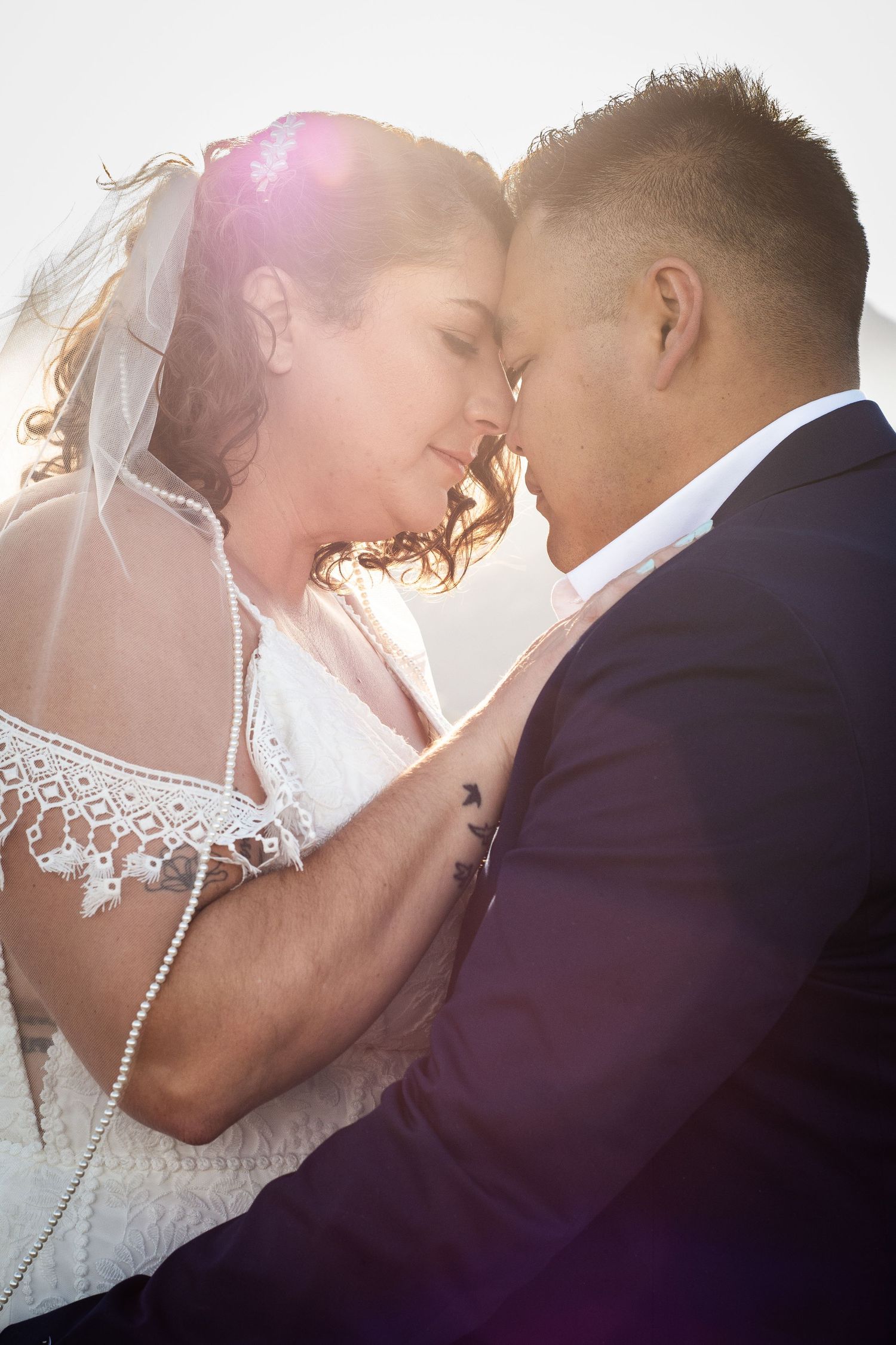 Bride and groom touching foreheads in tender romantic moment with bright backlight.