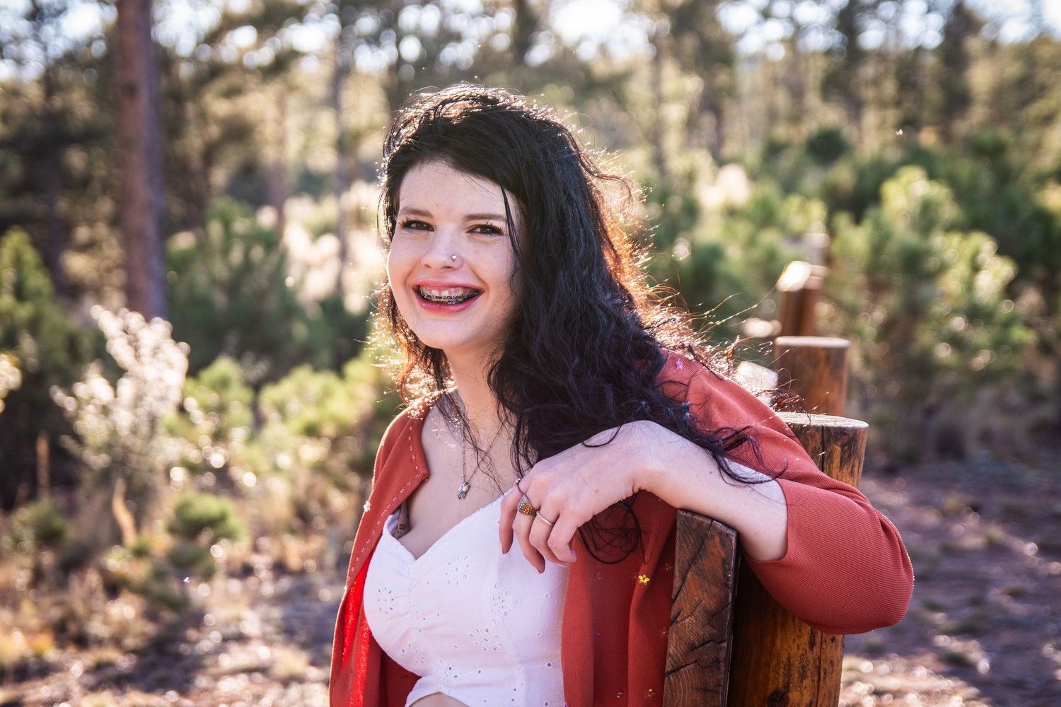 Smiling woman with dark curly hair in red jacket leaning on wooden post surrounded by pine trees outdoors.