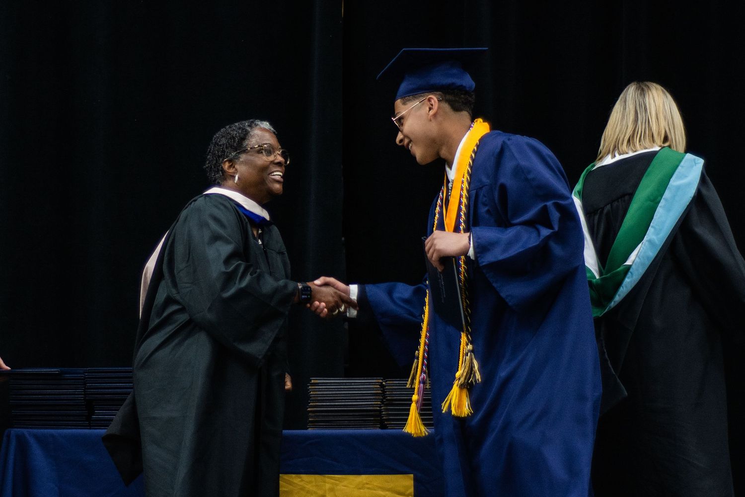 Graduate in navy cap and gown shakes hands with official in black robes at commencement ceremony.