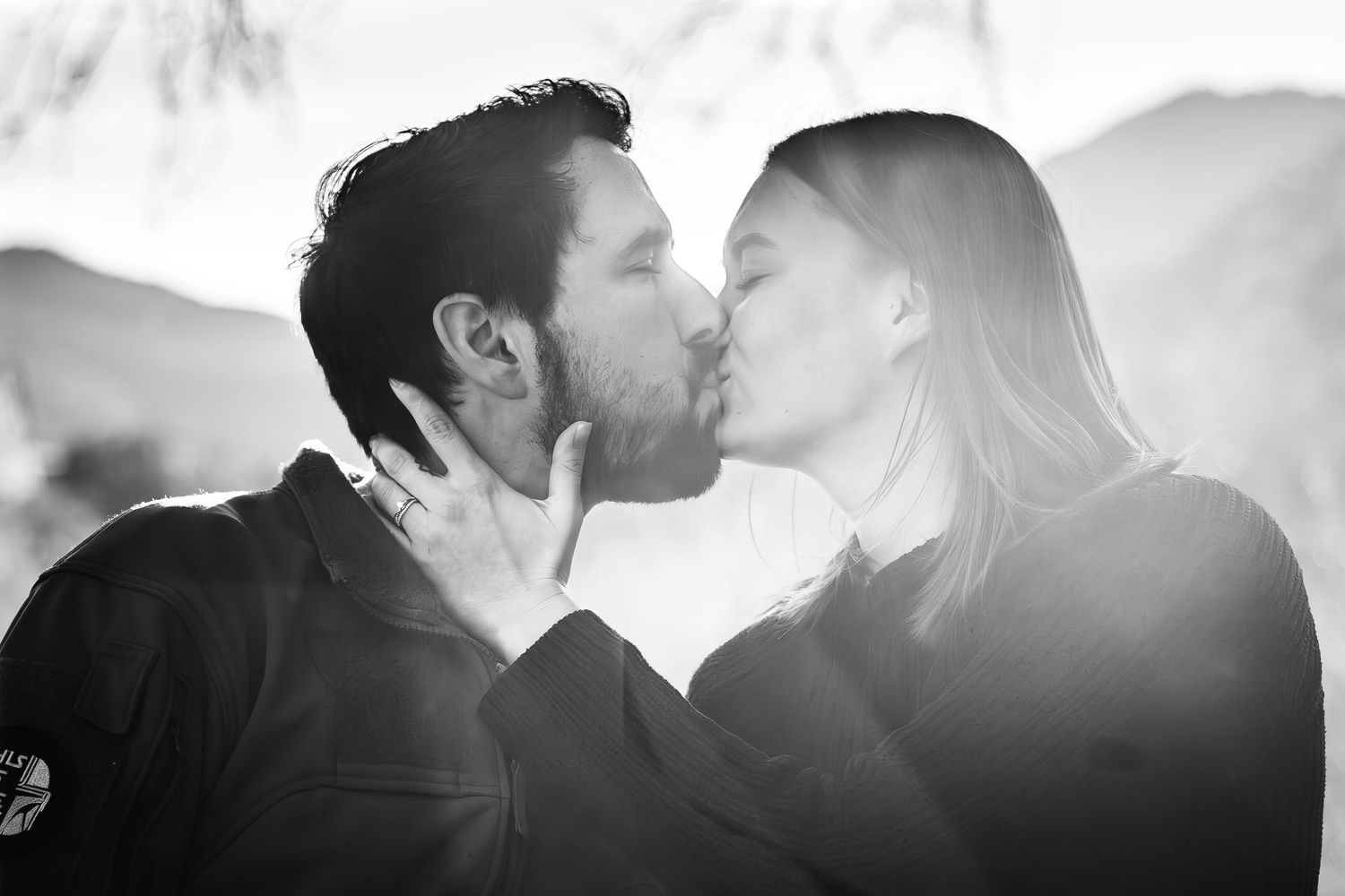 Black and white photo of a couple sharing a romantic kiss outdoors with soft backlit mountains in the background.