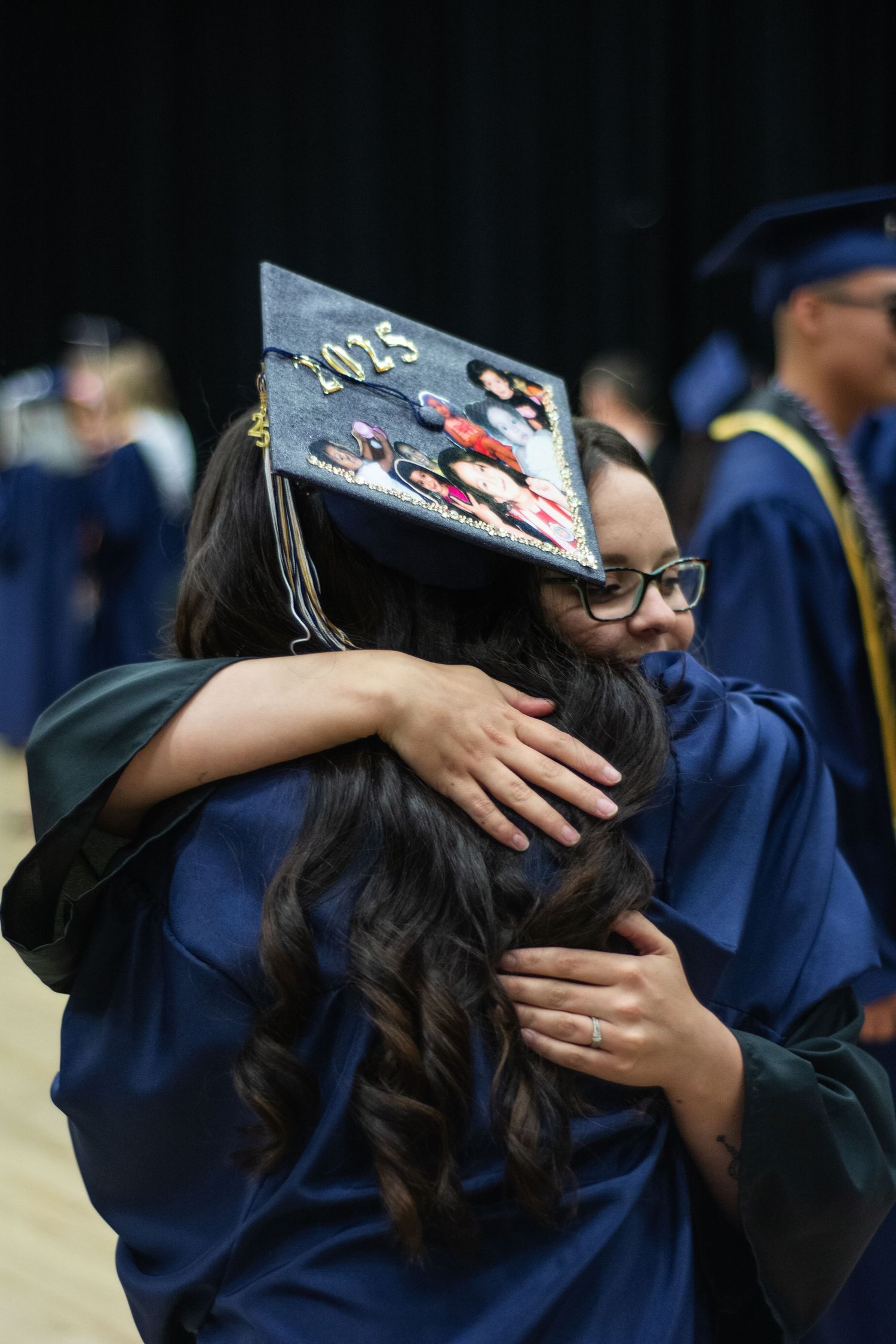 Two graduates in navy caps and gowns embrace warmly, one cap decorated with class of 2025 art.