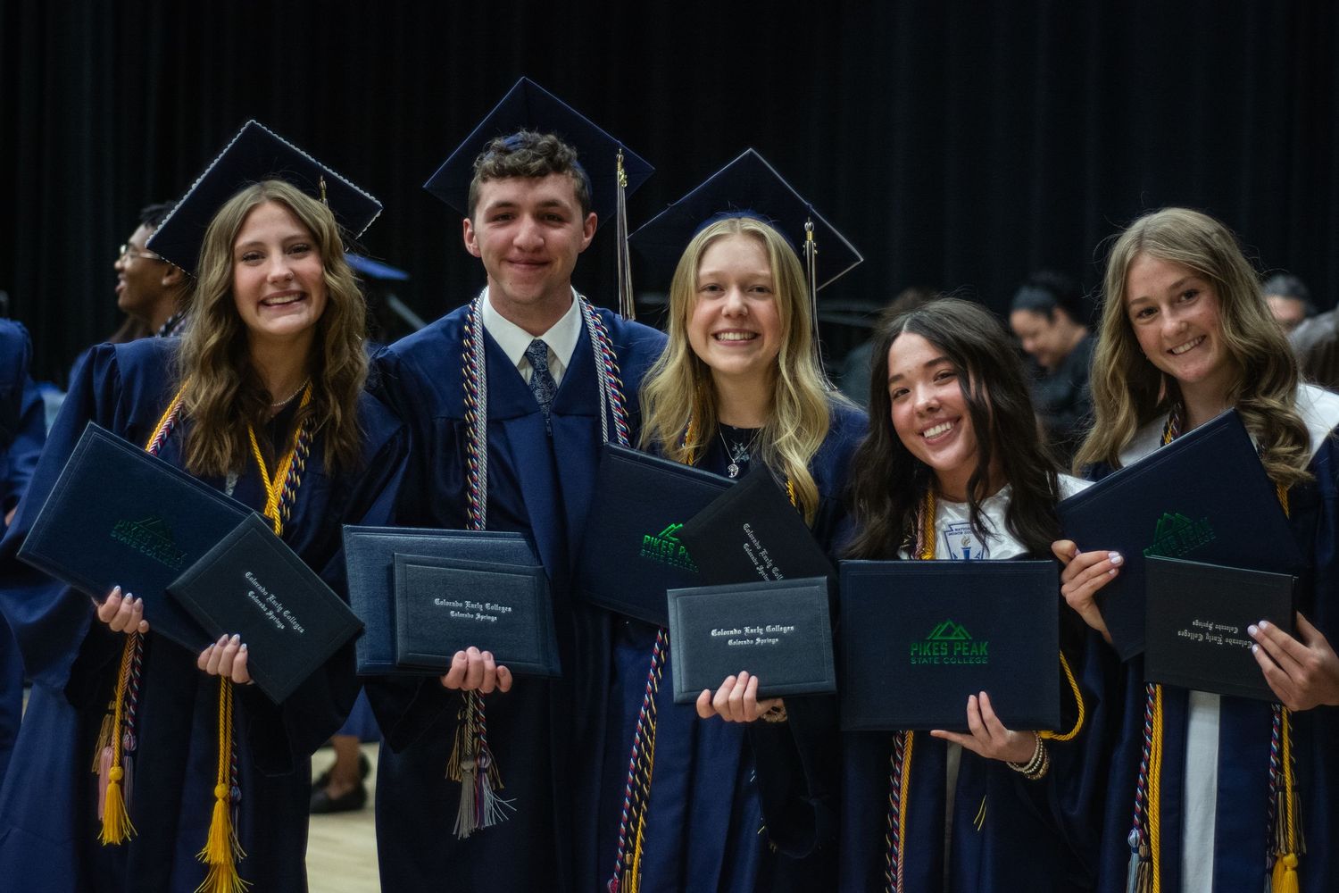 Five smiling graduates in blue caps and gowns proudly display diplomas at college commencement.