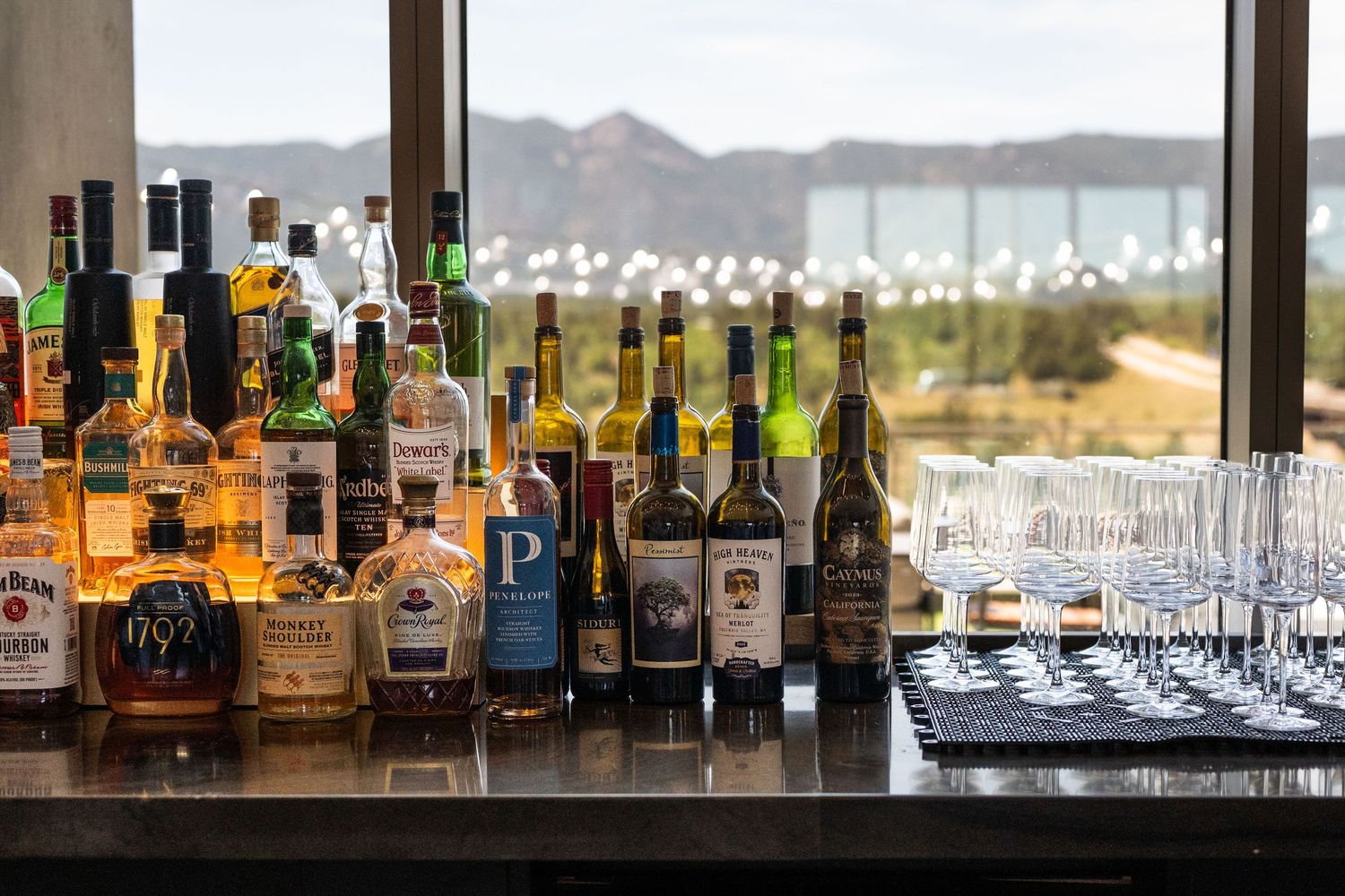 Assorted liquor bottles displayed on a bar counter with mountain views through large windows.