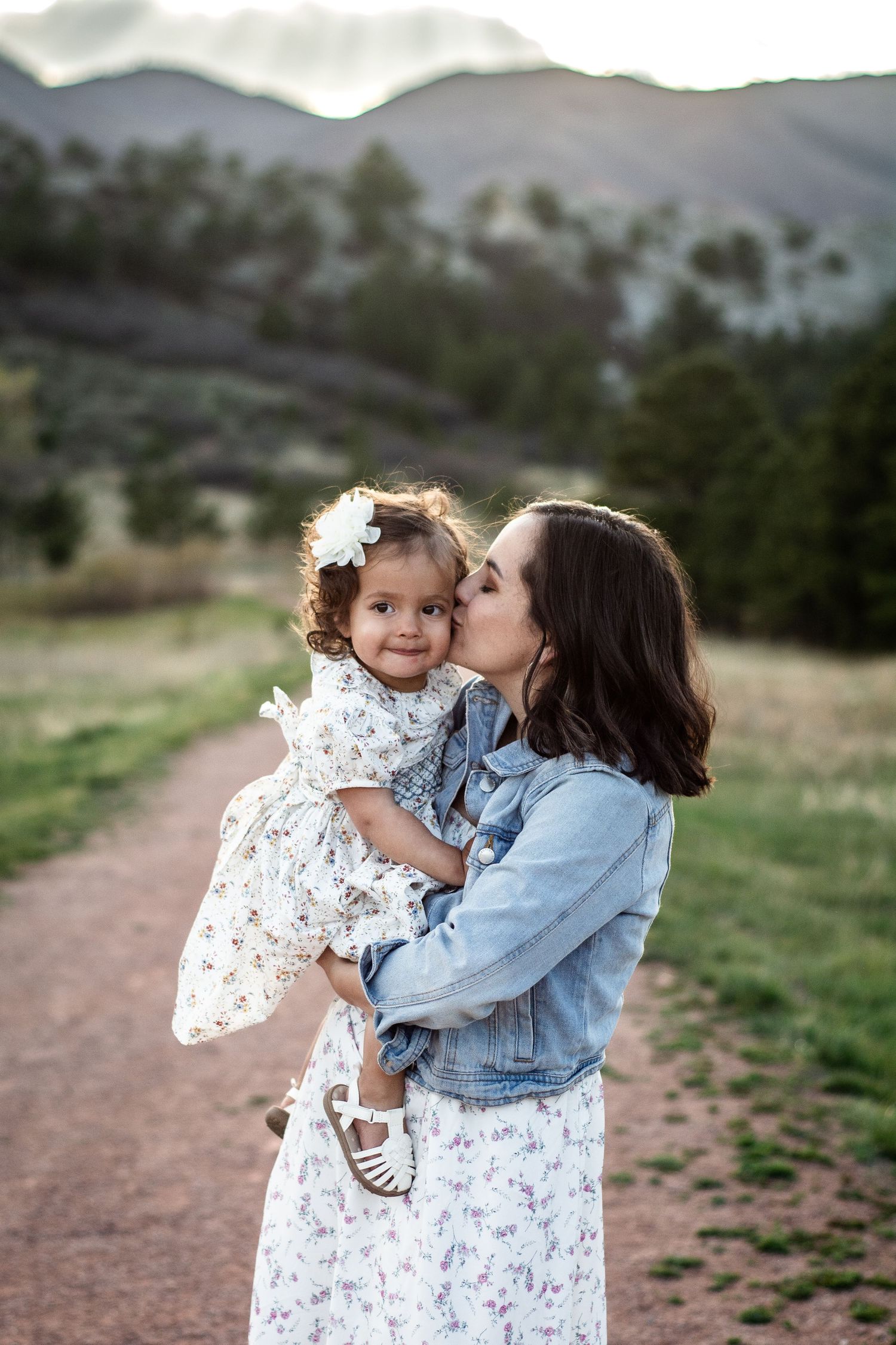 A mother in a floral dress and denim jacket kisses her toddler daughter outdoors with mountains in the background.