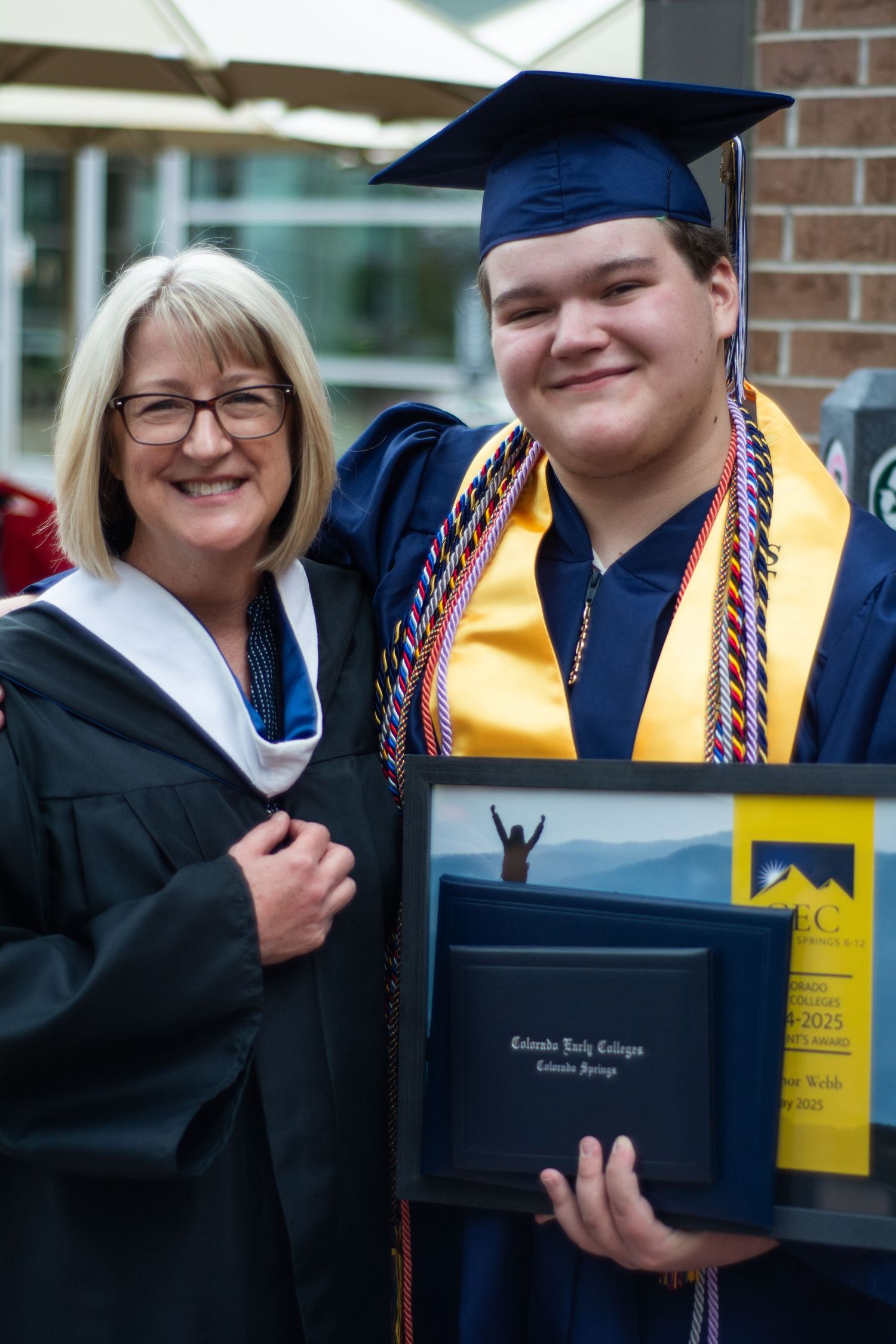 Graduate in blue cap and gown holds diploma at Colorado Early College commencement ceremony with proud faculty member.