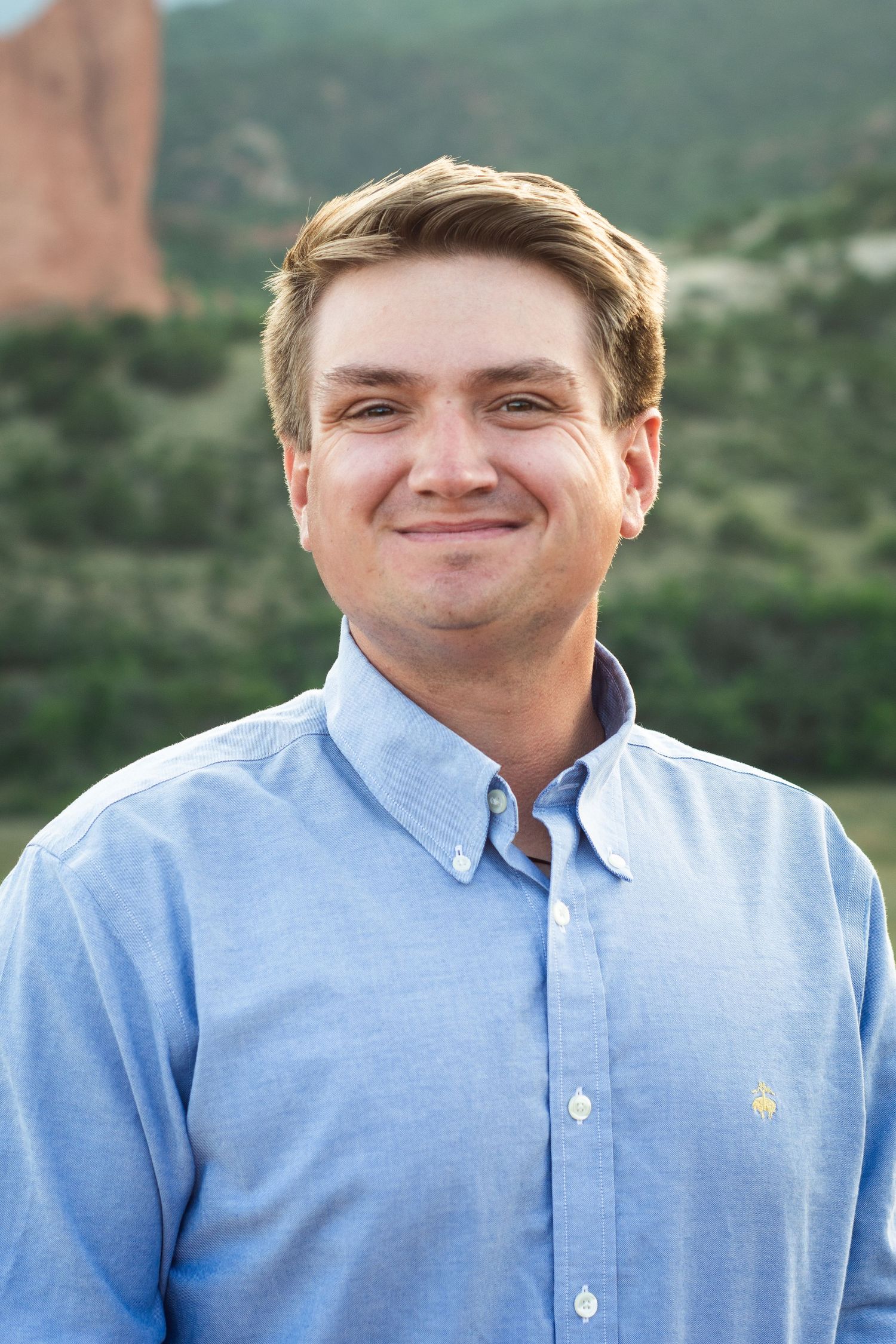 Smiling young man in blue button-down shirt posing outdoors with red rock formations behind him.