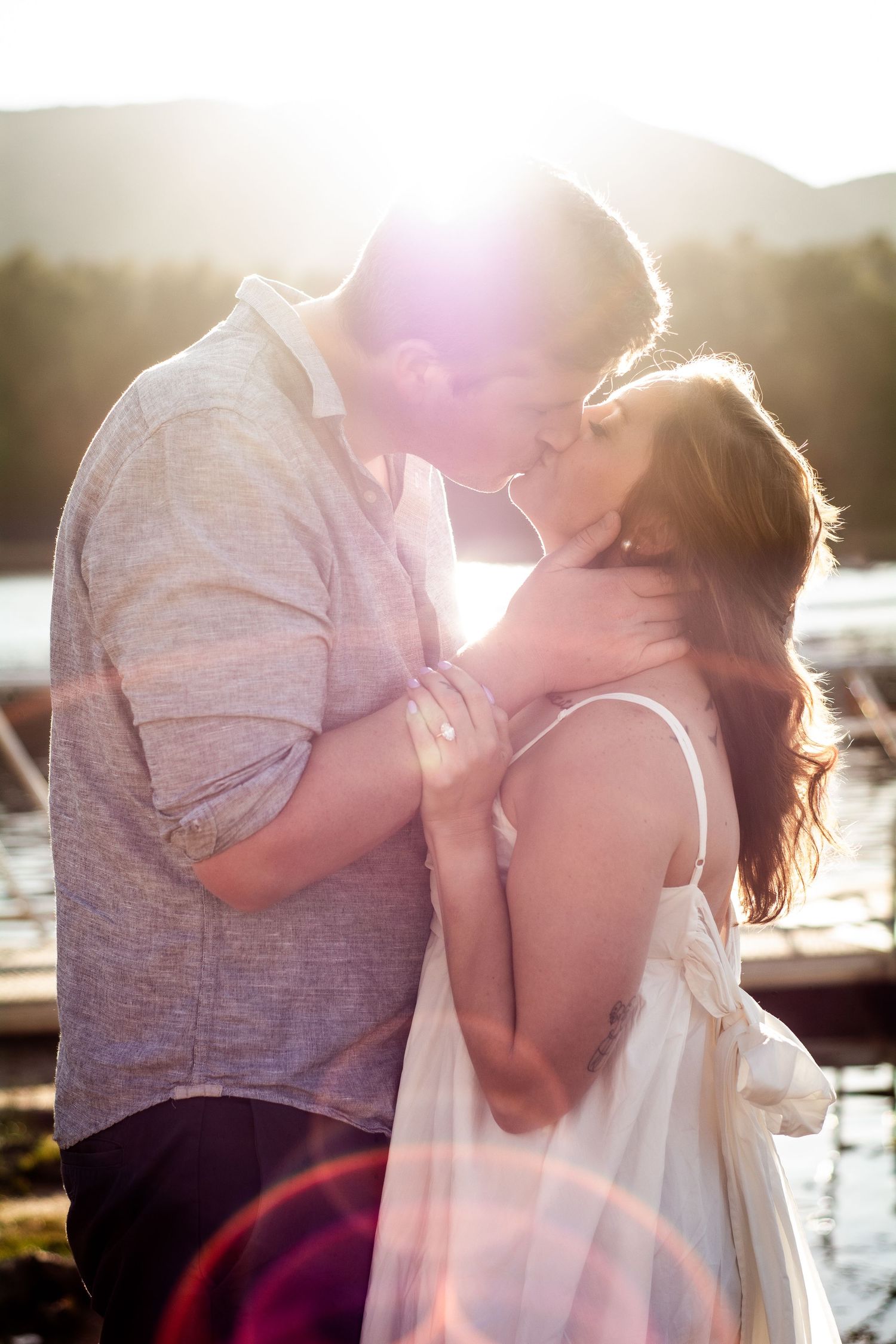Couple sharing a tender kiss backlit by golden sunset light near a lakeside dock.