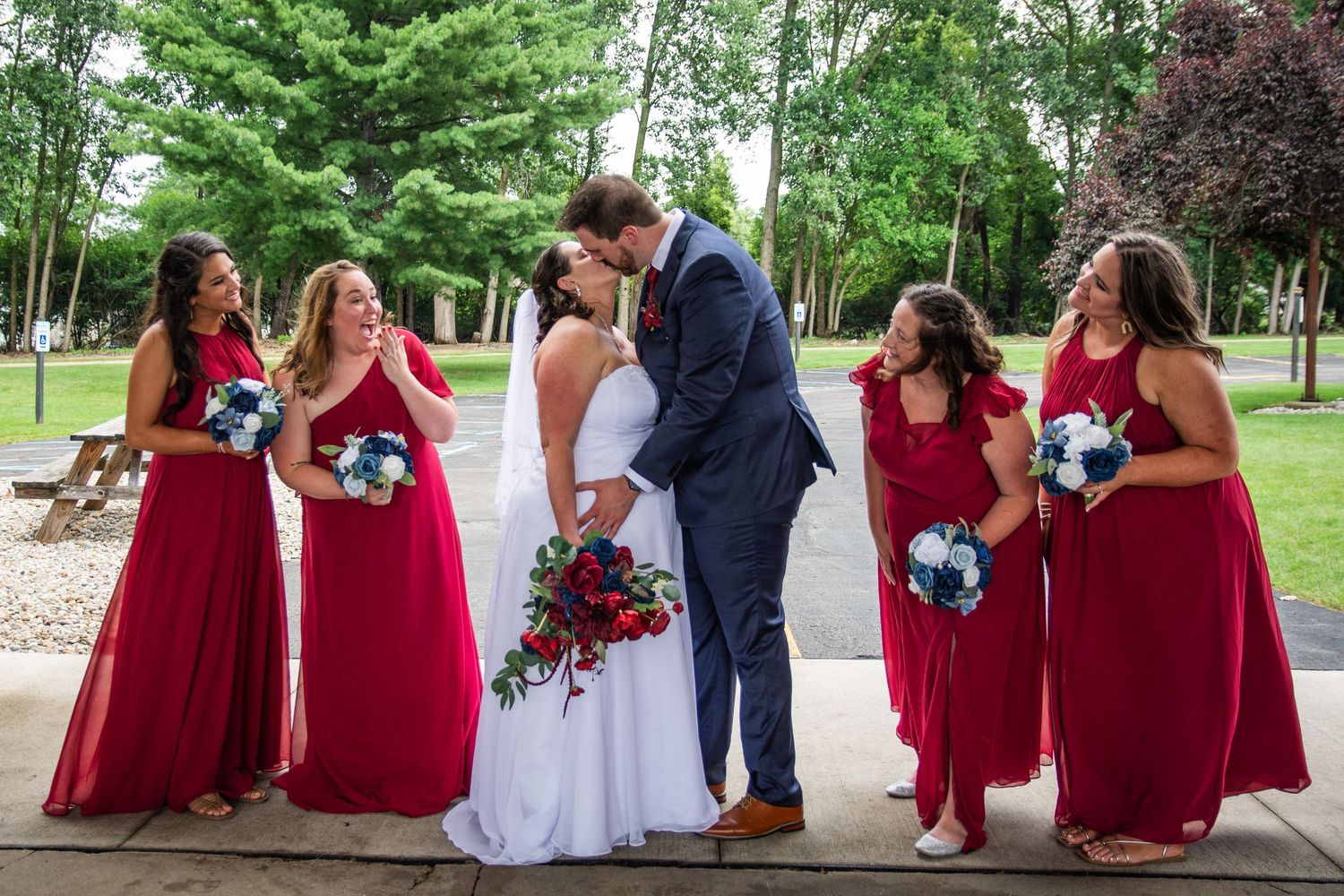 Bride and groom kissing surrounded by bridesmaids in red gowns holding bouquets outdoors at a wedding ceremony.