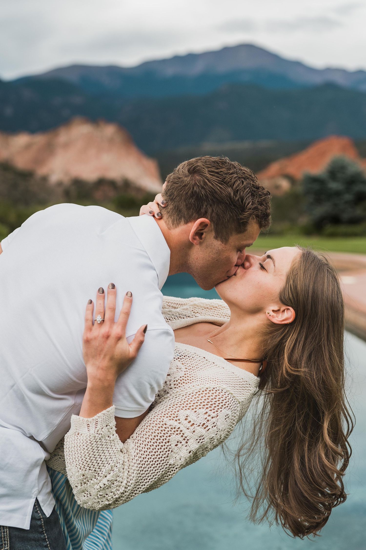 A couple shares a romantic dip kiss outdoors with mountain scenery and red rocks in the background.