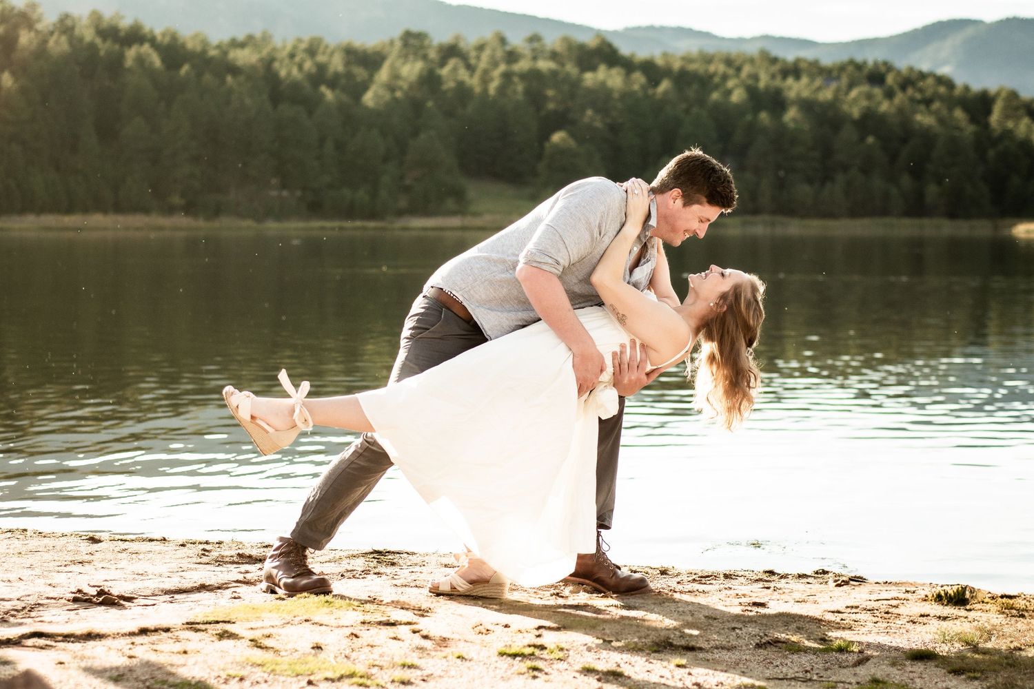 Romantic couple sharing a dip kiss by a serene lake, groom in gray suit, bride in white dress, mountains behind.