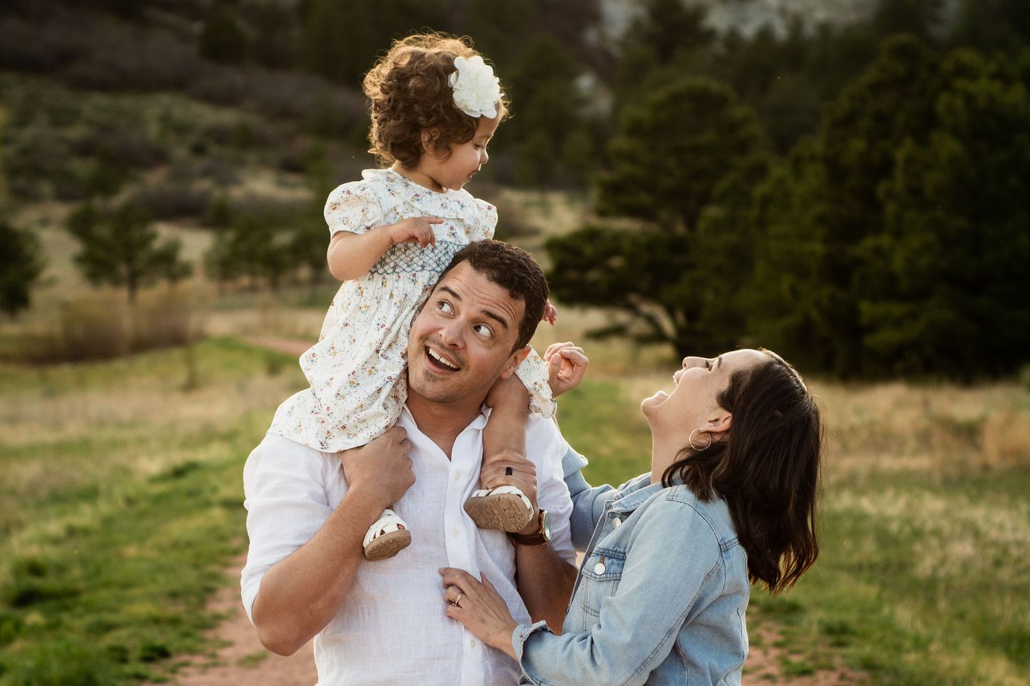 Happy family with toddler girl on dad's shoulders laughing outdoors in a green meadow during a portrait session.