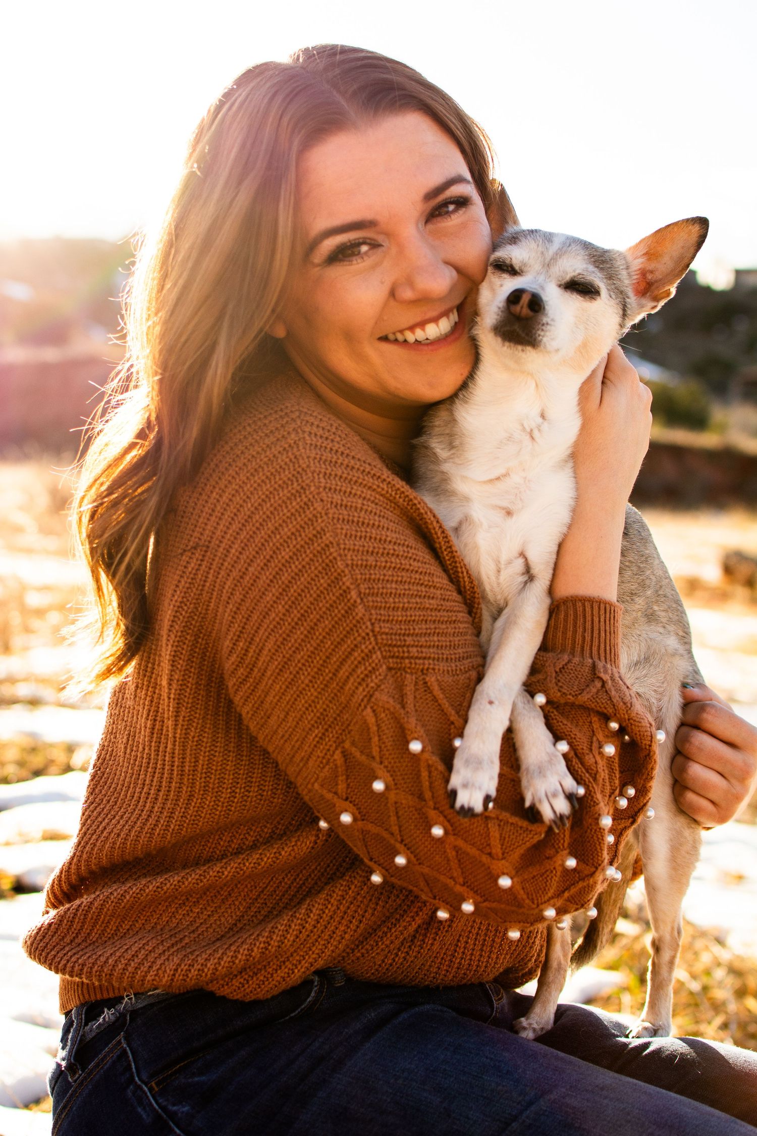 Smiling young woman in brown sweater hugging a small Chihuahua dog outdoors in golden sunset light.