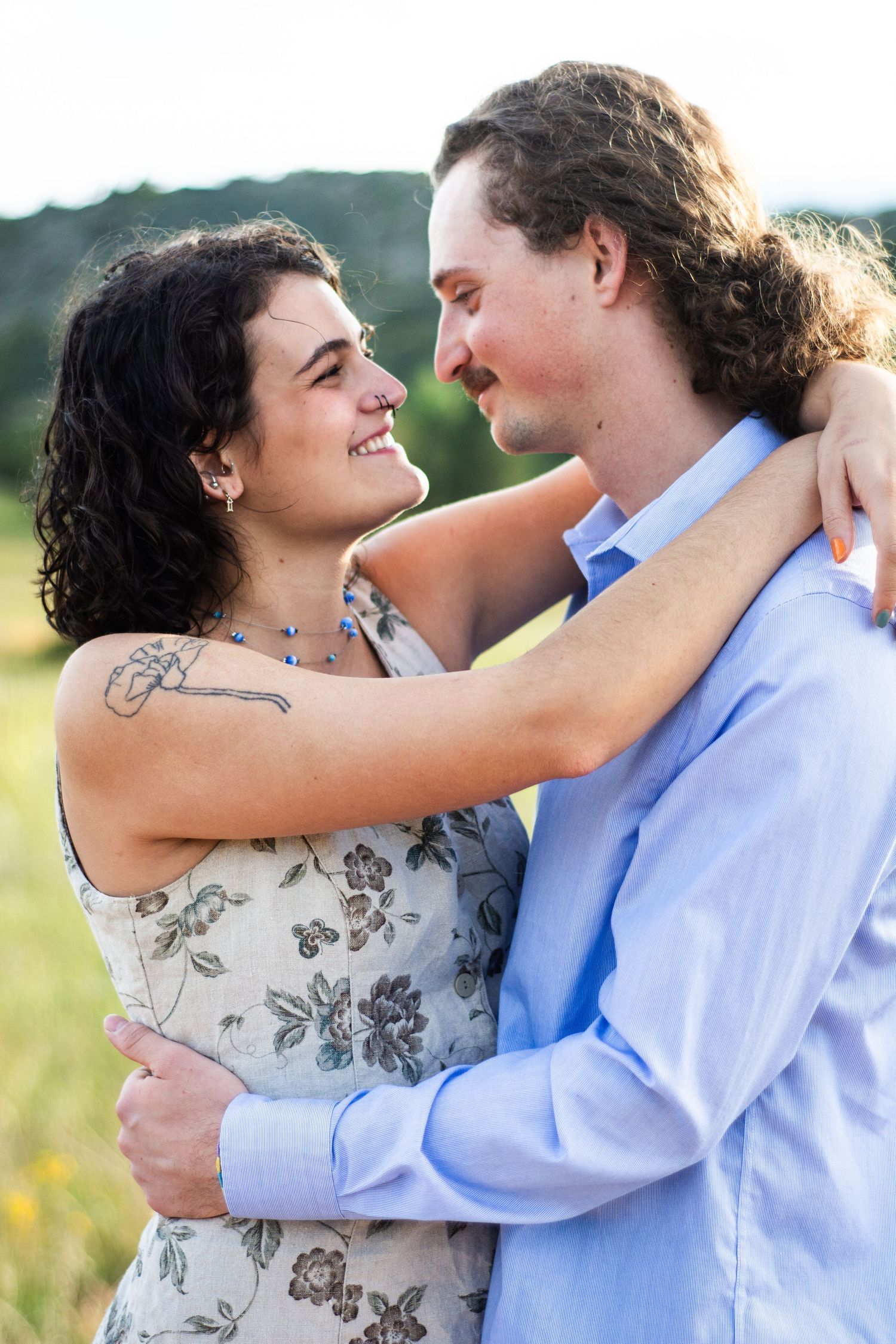 A smiling couple embraces outdoors, the woman in a floral dress with a tattoo, the man in a light blue shirt.