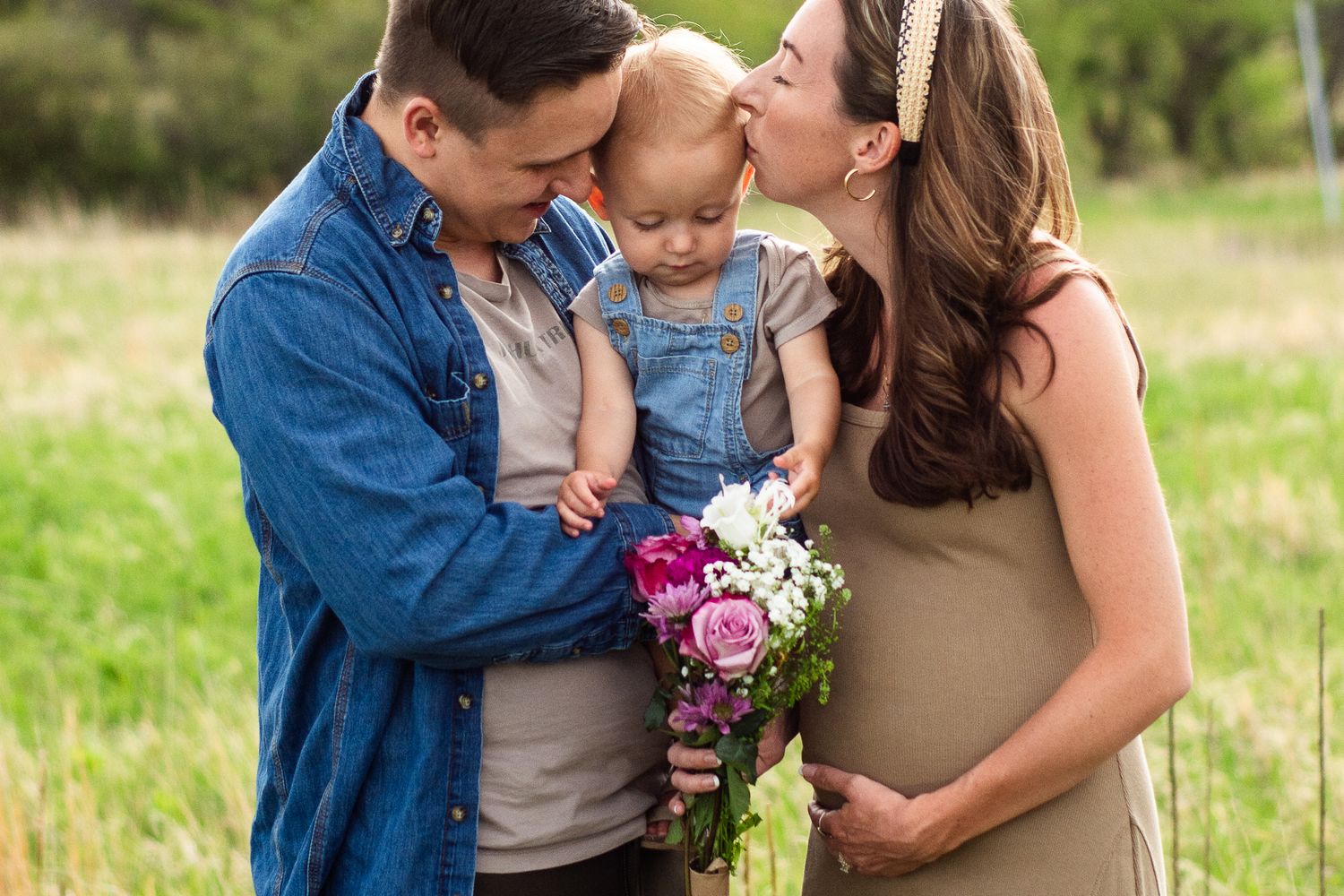 Pregnant woman and man holding toddler kissing cheek outdoors in field with pink and white flower bouquet.