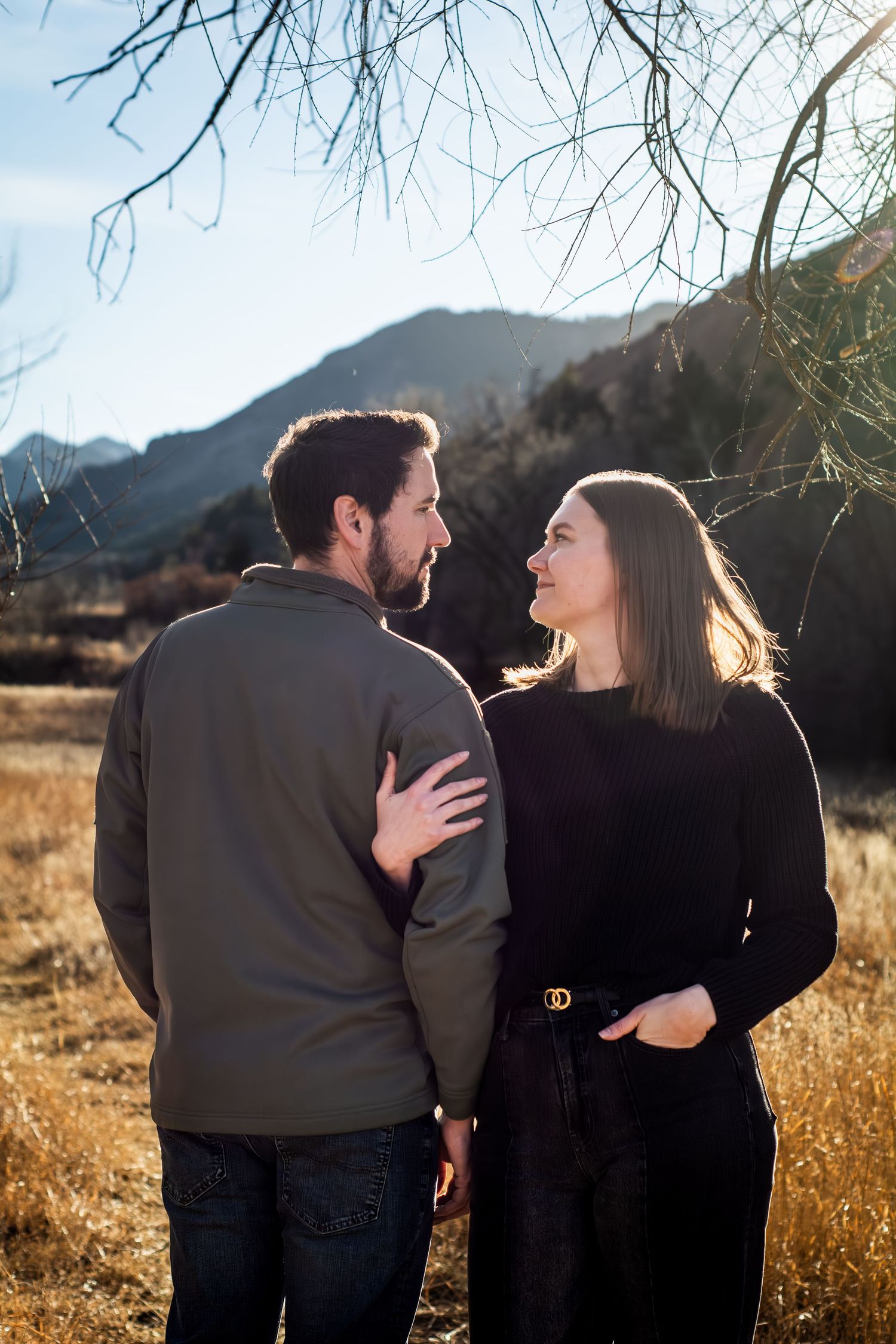 Couple sharing a romantic moment in a mountain meadow, woman in black sweater and man in olive jacket embracing outdoors.