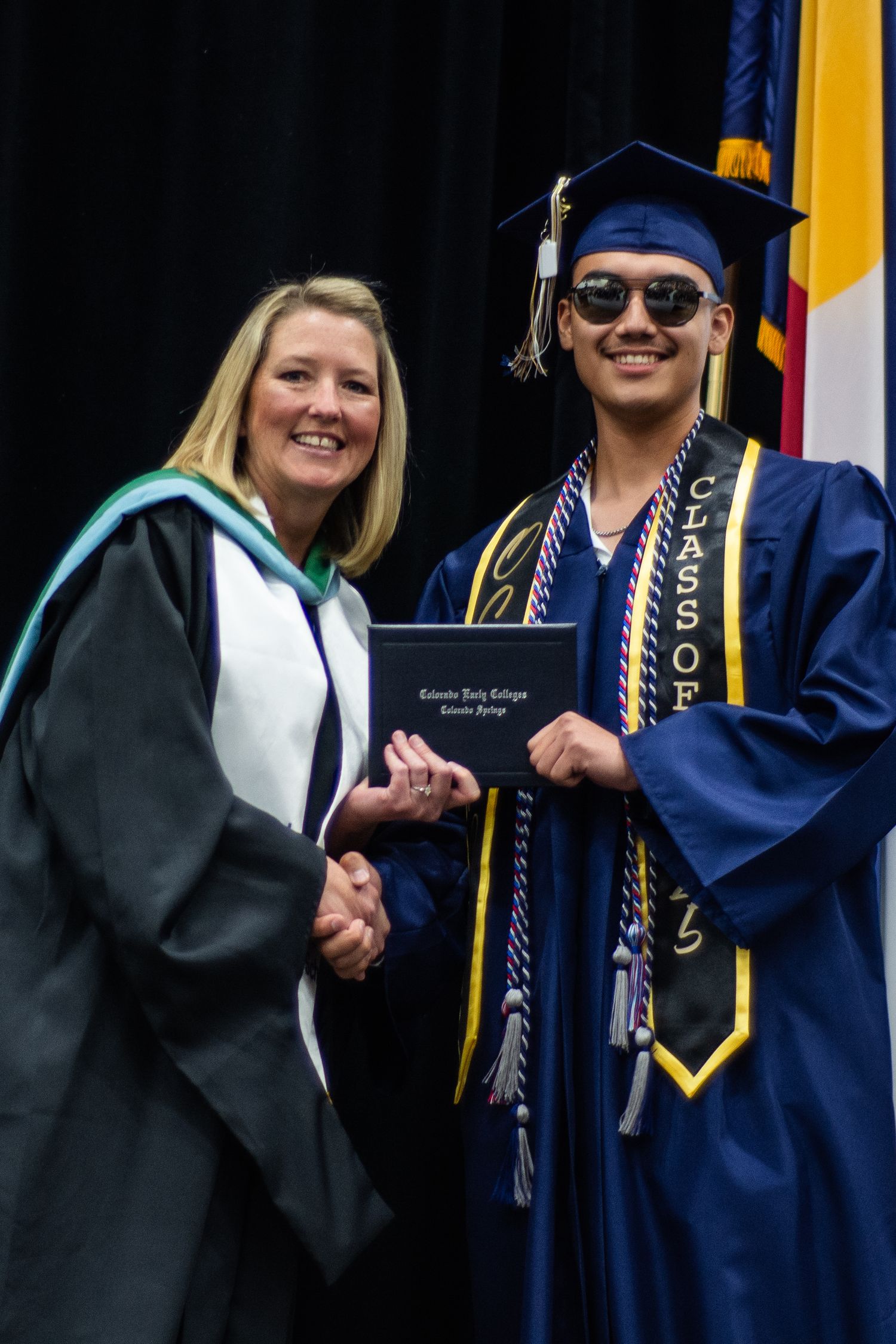 Smiling graduate in sunglasses receives diploma from administrator at college graduation ceremony.