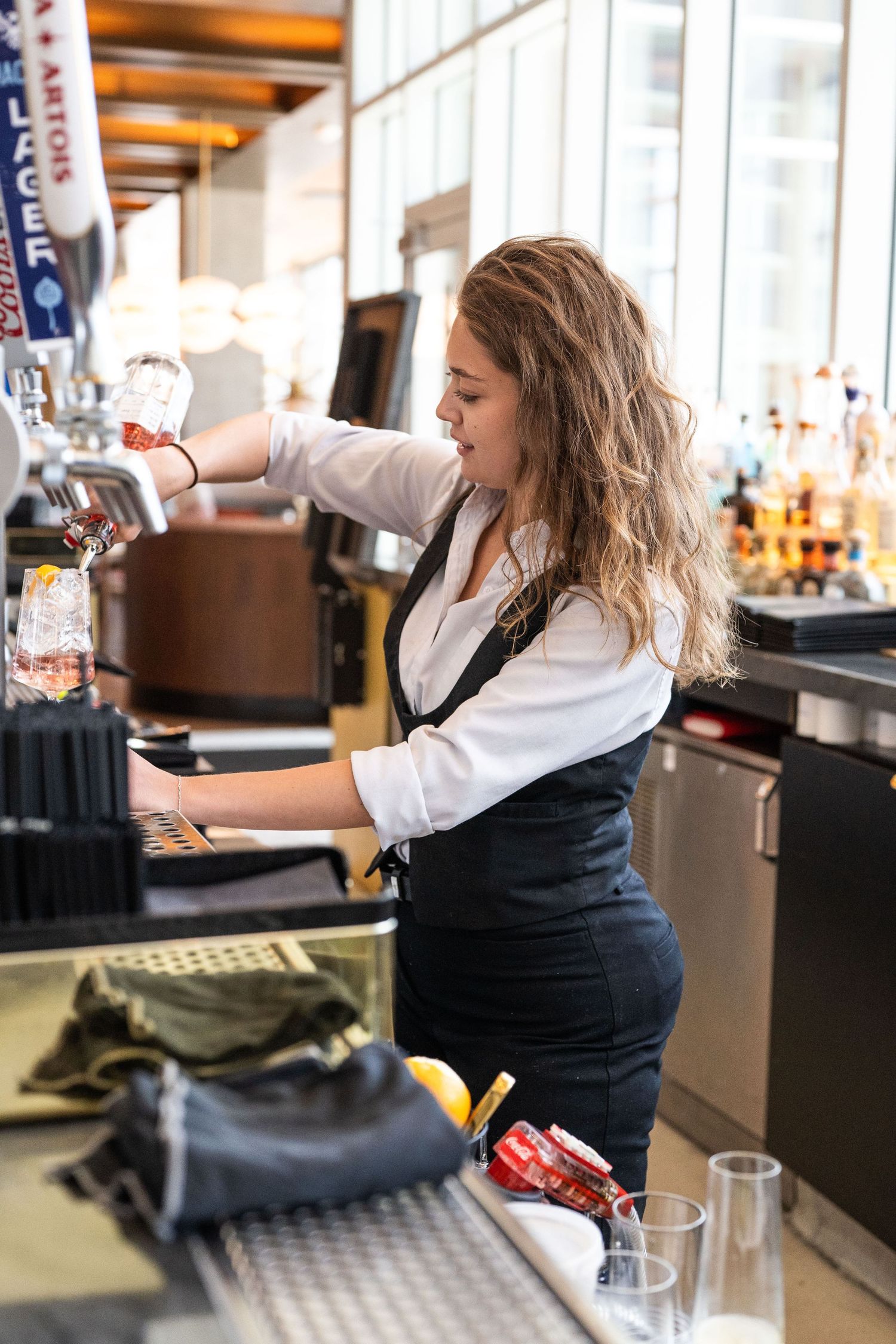 Female bartender with curly hair pouring draft beer at a modern bar counter in a white shirt and vest.