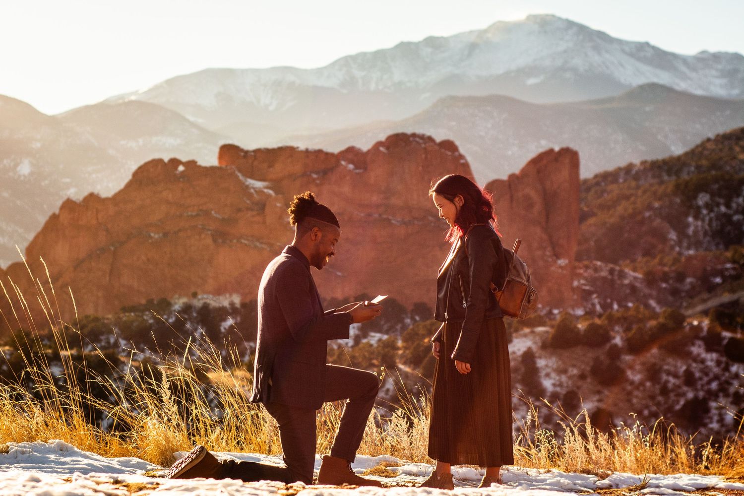 Man proposing to woman on one knee at Garden of the Gods, Colorado, with red rock formations and mountains at sunset.