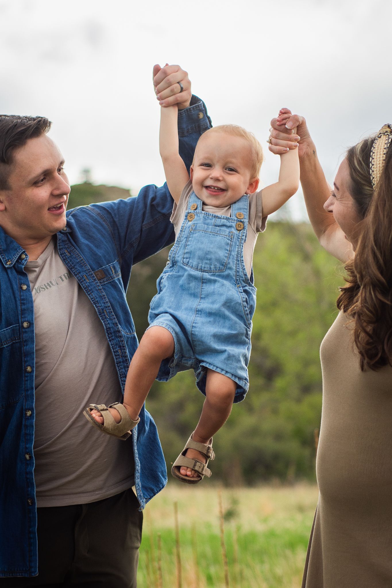 Happy baby in denim overalls lifted by smiling parents outdoors in a green field.