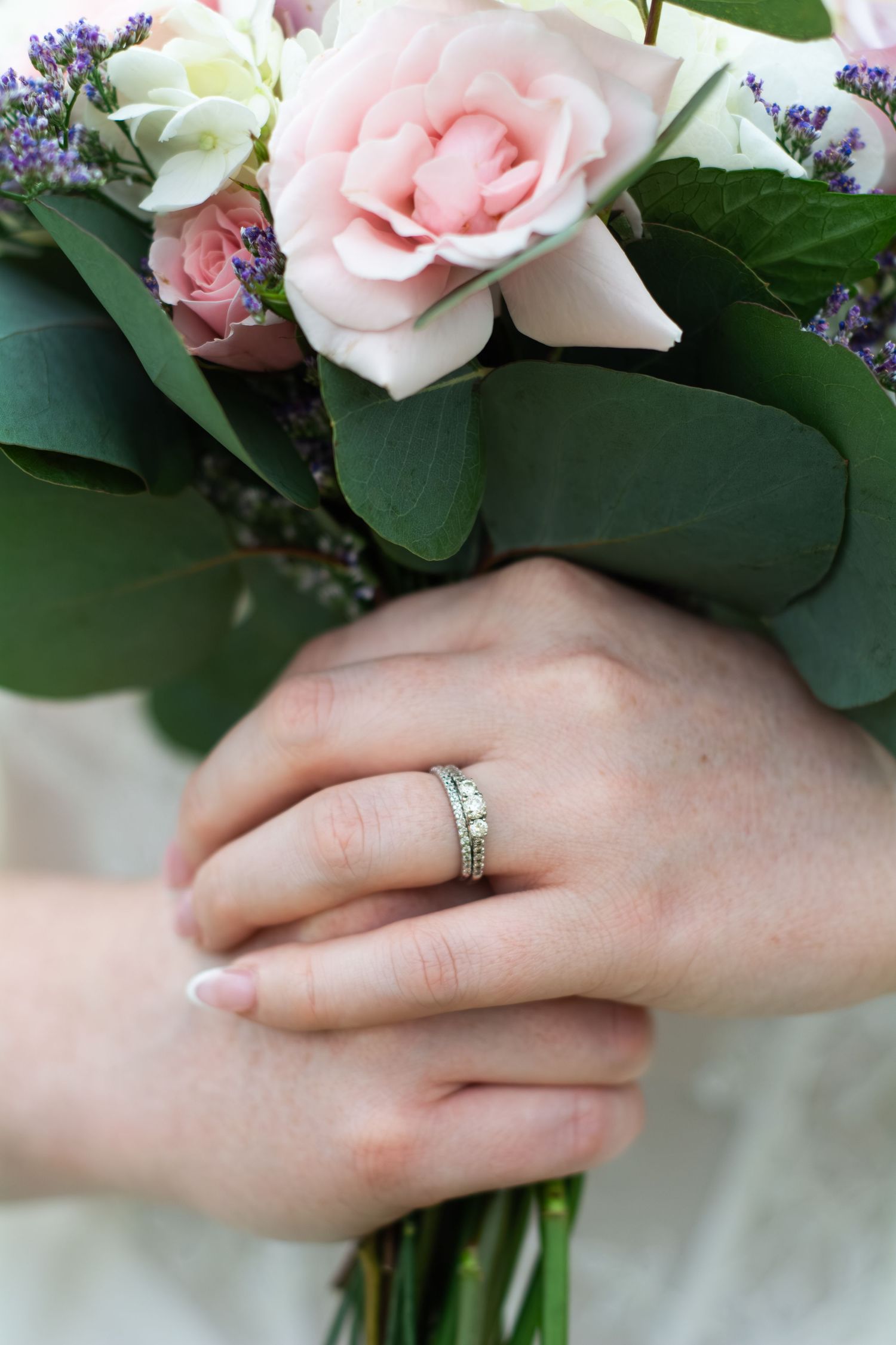 Bride holding pink rose bouquet with greenery, showcasing diamond wedding ring set on manicured hand.