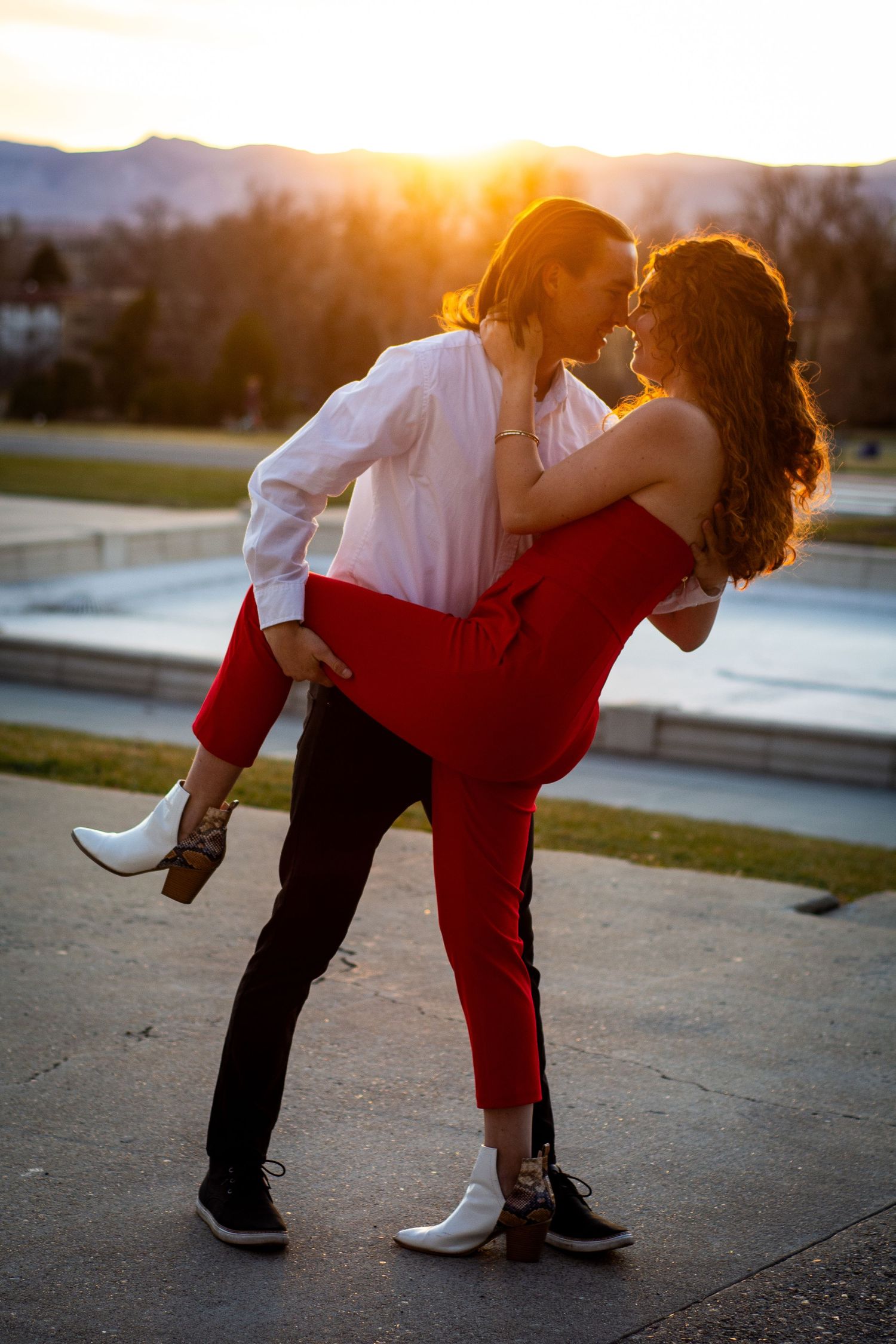 Couple sharing a romantic kiss at sunset, man lifting woman in matching red outfits with golden backlight.