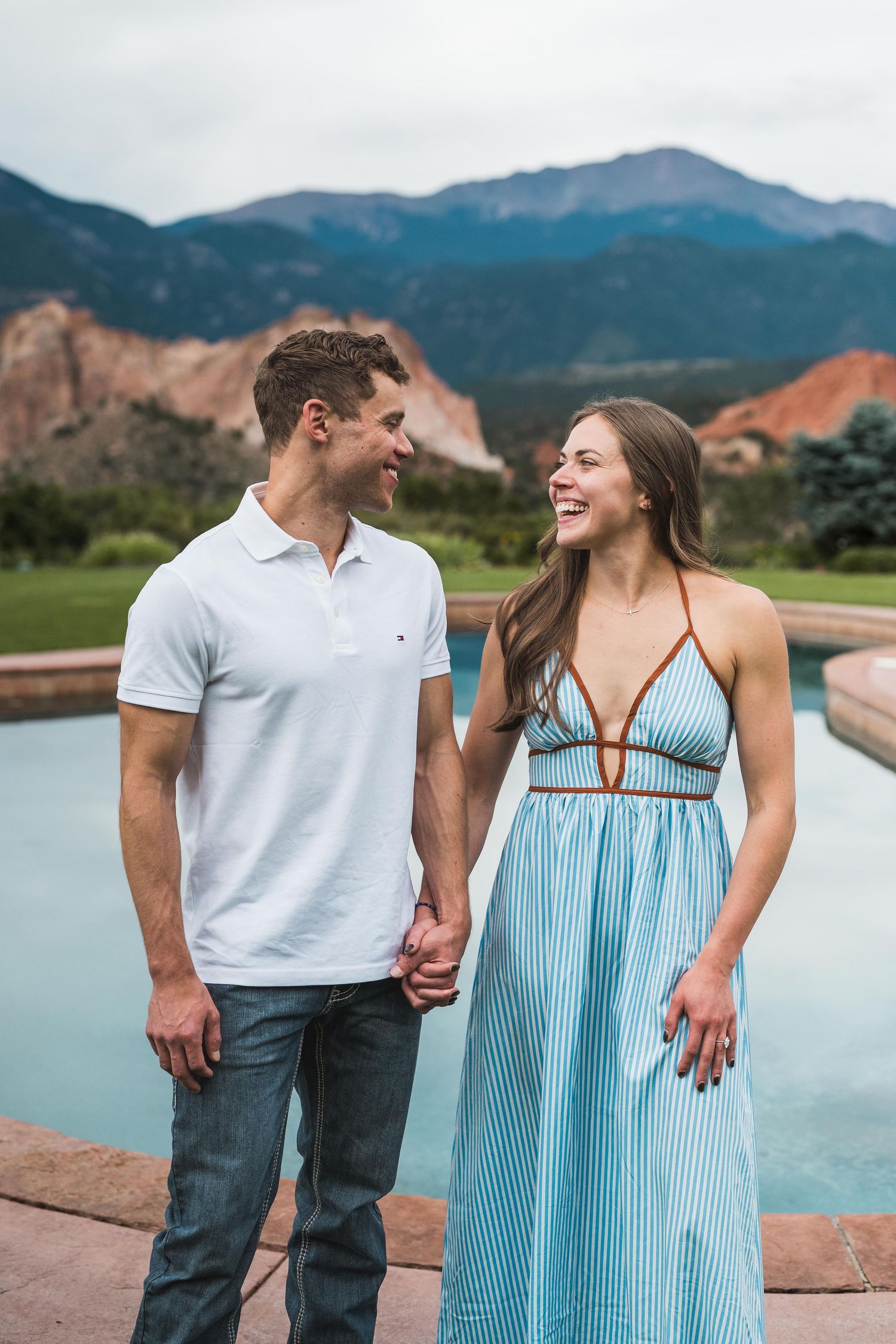 A smiling couple holds hands by a pool with red rock mountains in the background on a cloudy day.