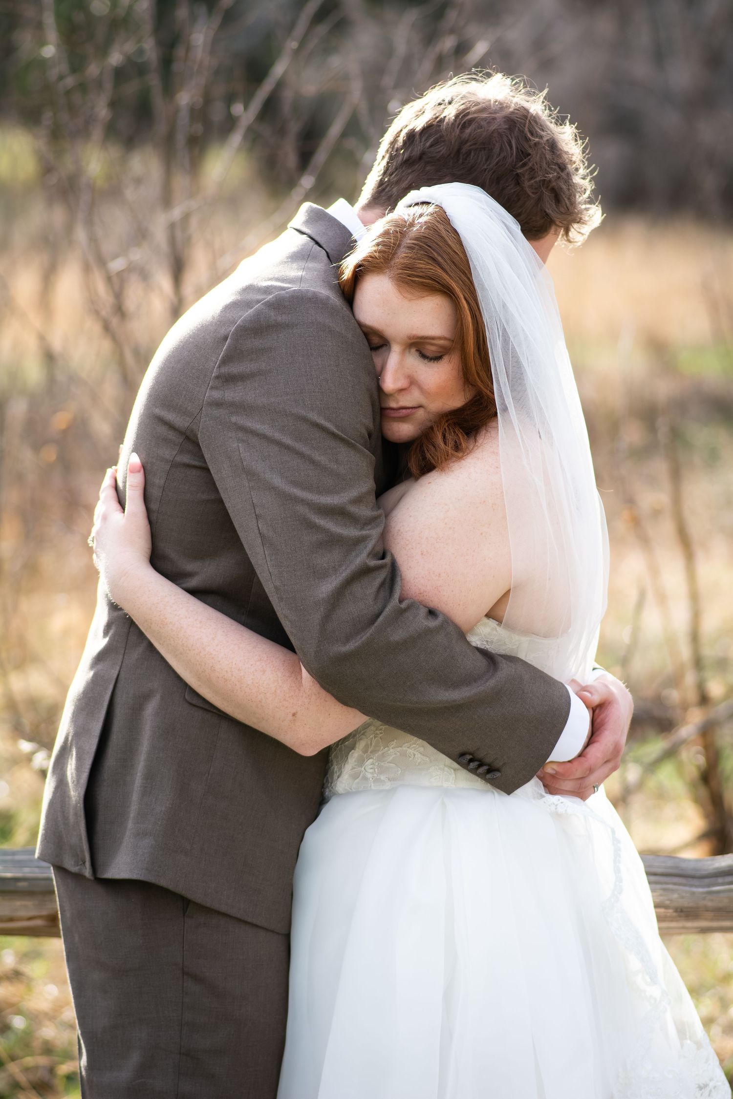 A bride and groom share a tender embrace outdoors, with the bride in a white gown and veil resting her head on his shoulder.