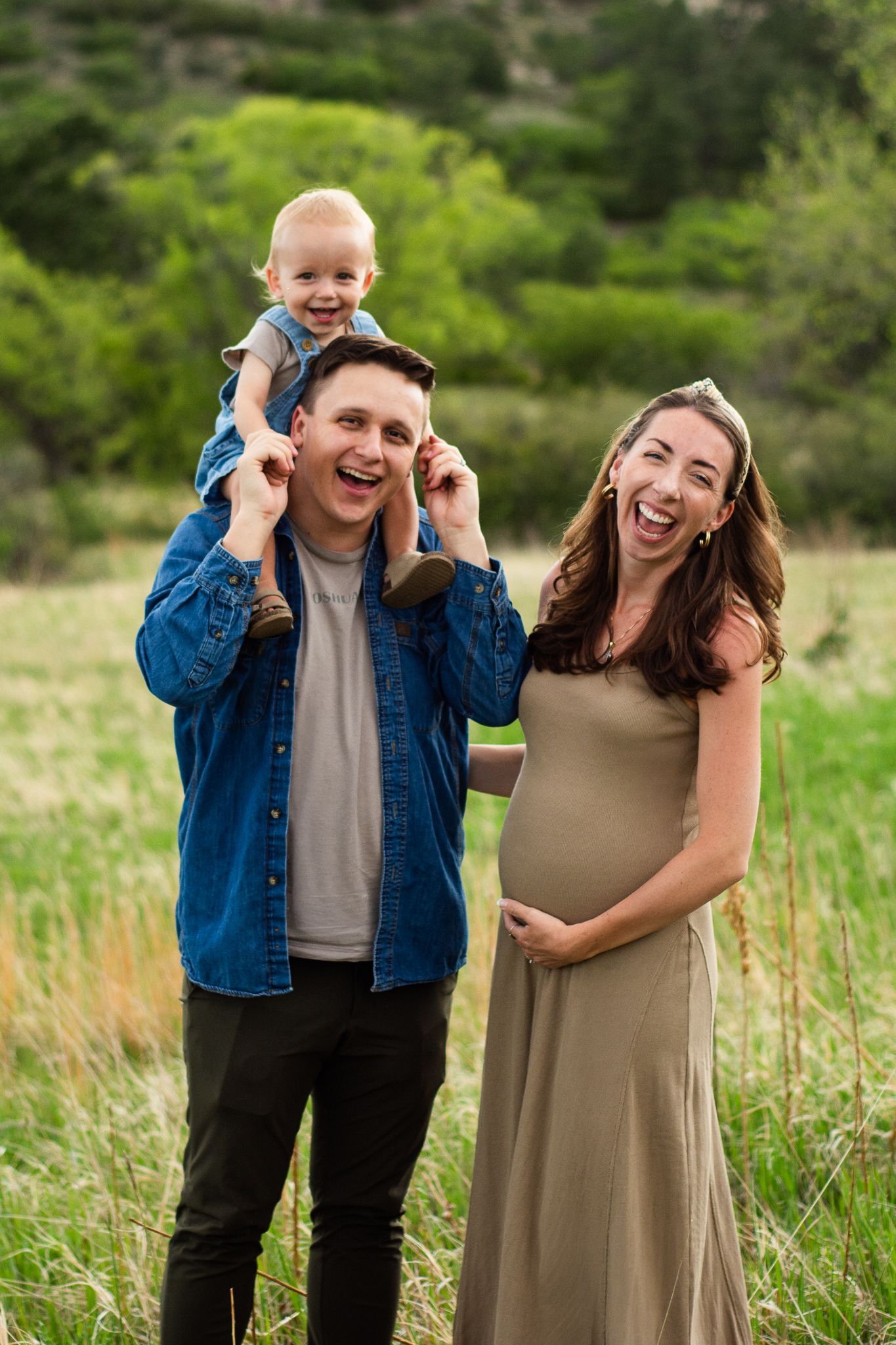 Happy pregnant family with toddler on dad's shoulders laughing in a green field during maternity photo session.