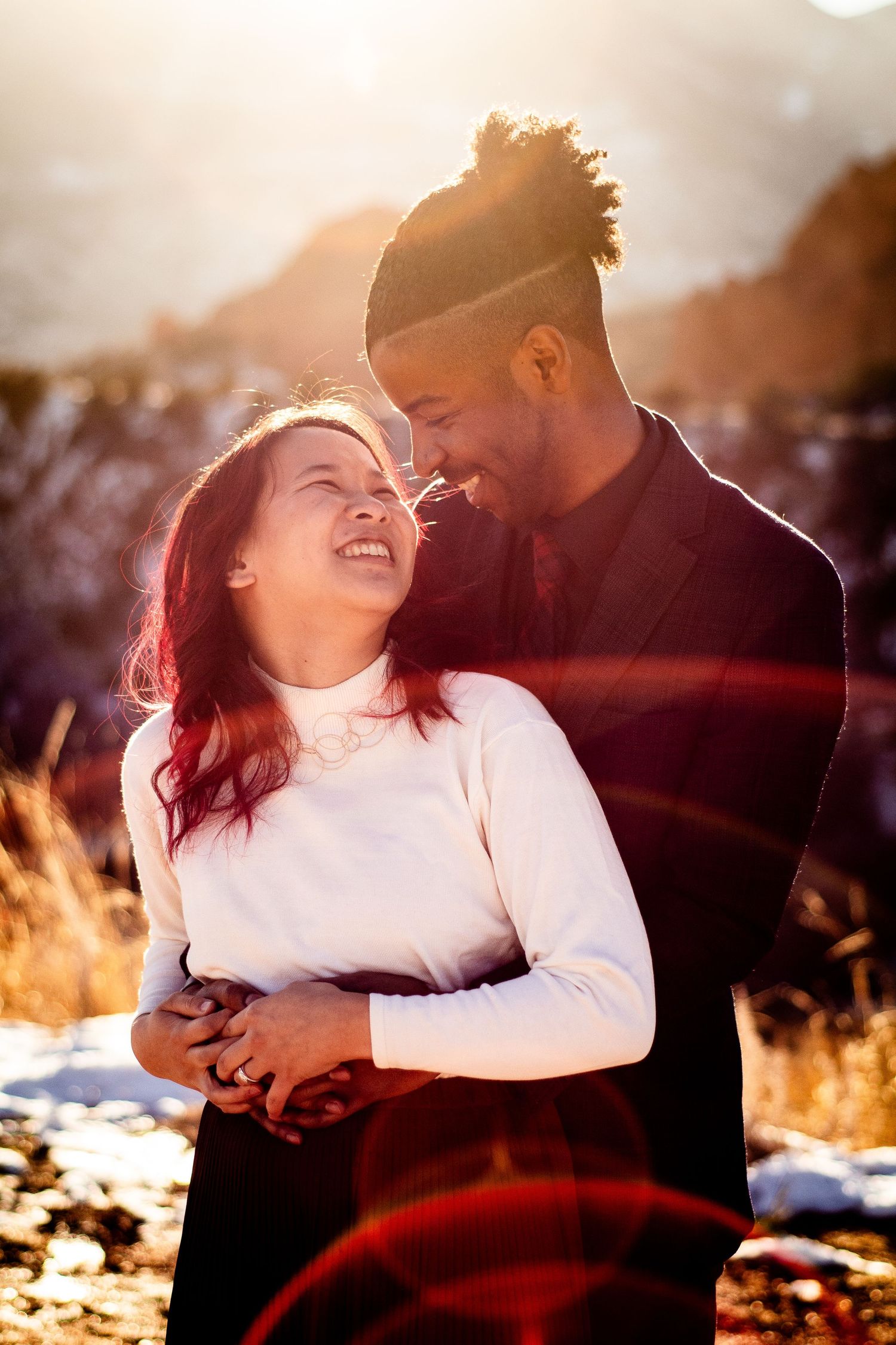 Couple embracing outdoors in winter sunlight, woman with red hair laughing joyfully as man smiles down at her.