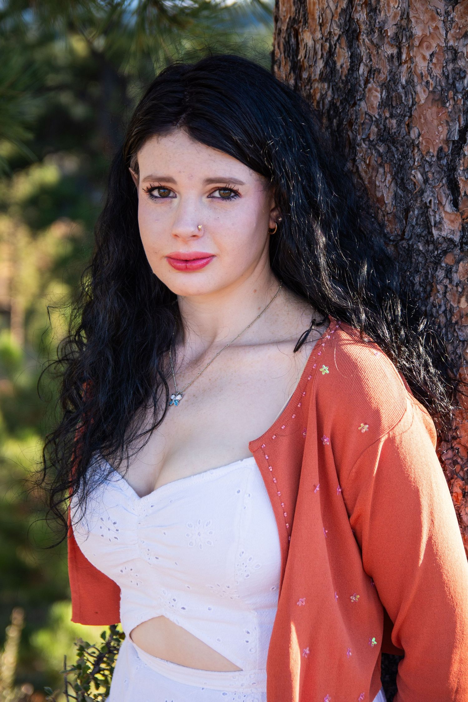 Young woman with dark curly hair wearing a white top and orange cardigan, leaning against a tree outdoors.