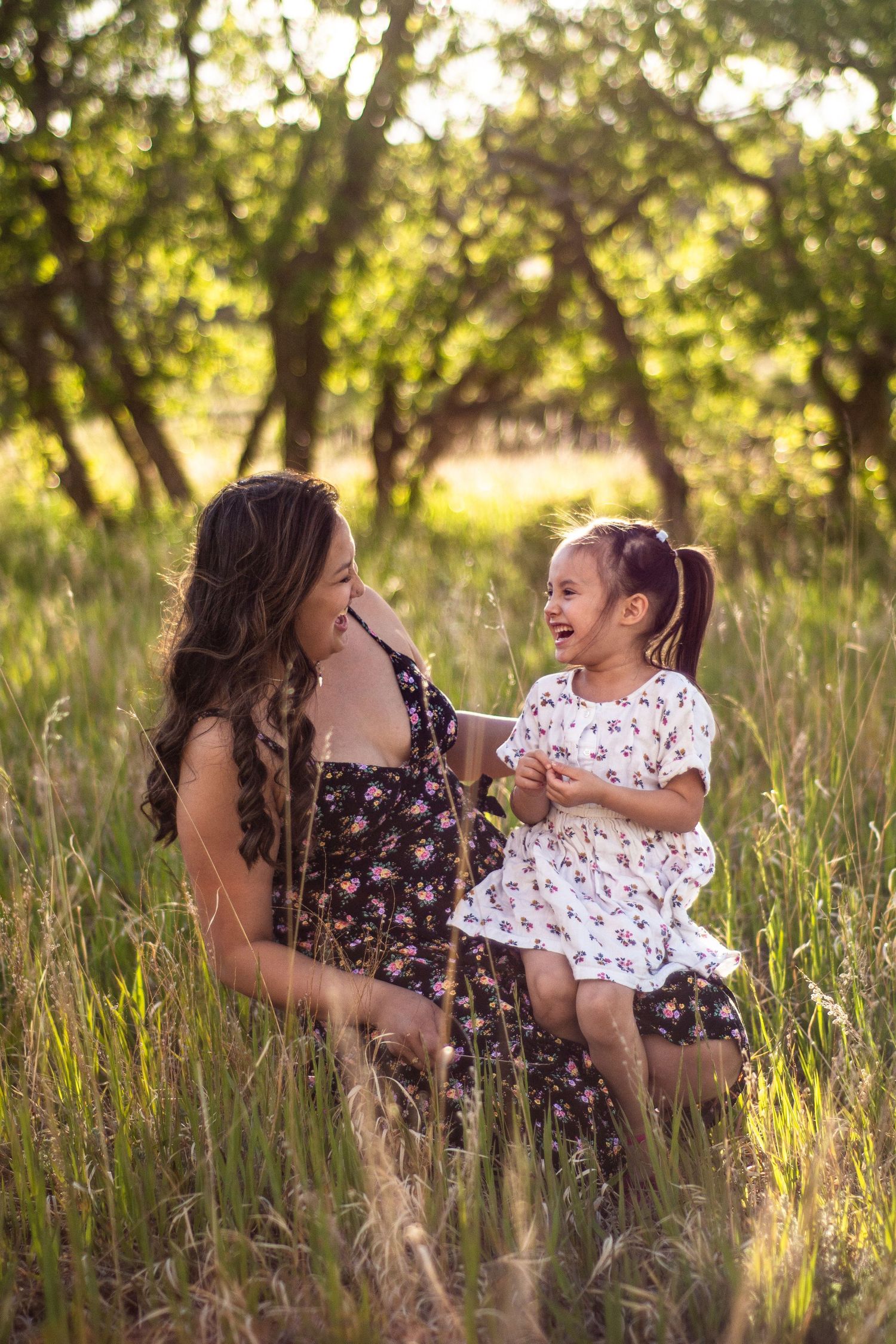 Mother in floral dress sits in tall grass with laughing toddler daughter in sunlit orchard setting.